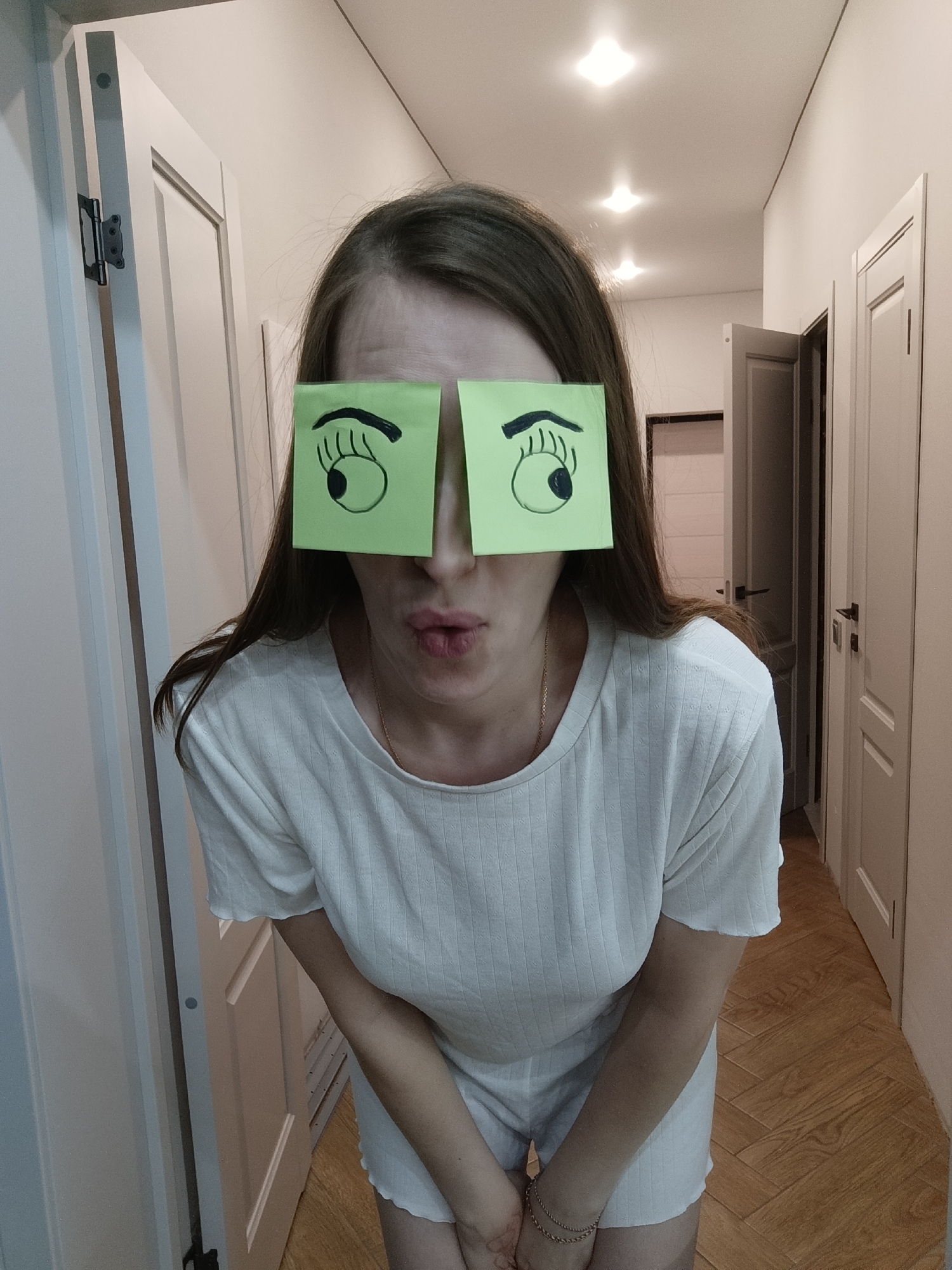 A woman with light brown hair has two green sticky notes with drawn eyes covering her eyes. She is wearing a white, ribbed t-shirt and light-colored shorts. Her arms are bent, and her hands are clasped in front of her waist. Her lips are puckered. She is standing in a hallway with light wood flooring. There are multiple white doors along both sides of the hallway. The door on the right side of the frame is slightly ajar. There are four light fixtures on the ceiling. The picture was taken in Chaikovsky, Russia.