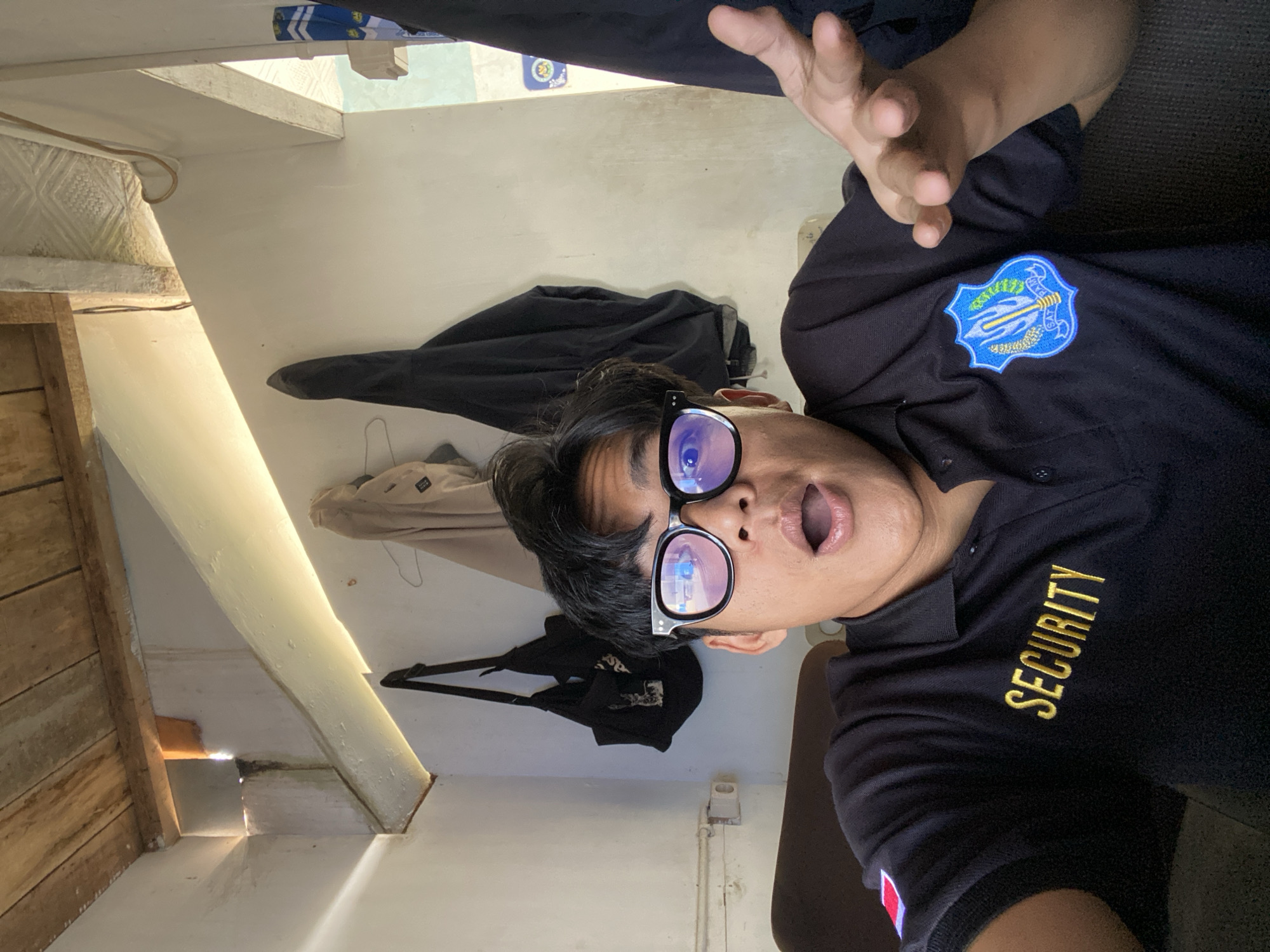 A young man wearing a black polo shirt with "SECURITY" embroidered in gold letters and a blue emblem on the chest takes a selfie. He has black hair and is wearing black-rimmed glasses. His mouth is open in surprise, and his right hand is raised with fingers spread. In the background, two black bags hang on the wall, along with a light-colored jacket. The room has a wooden ceiling and white walls, with light streaming in from an opening. The man appears to be inside a building.
