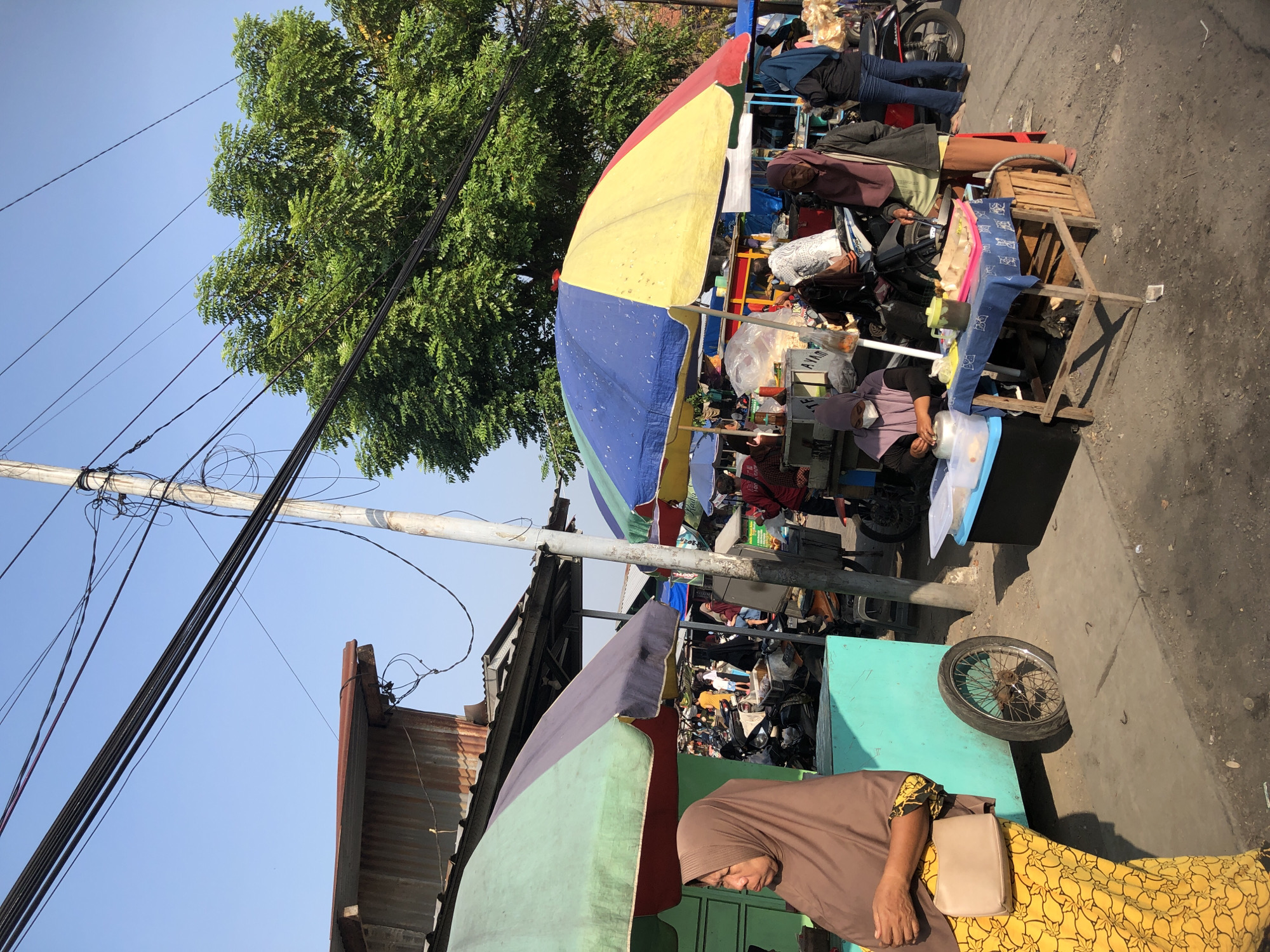 In Balongdowo, Indonesia, a bustling street market unfolds under a clear blue sky. The foreground features a woman in a brown hijab and a yellow patterned dress, carrying a beige purse. Adjacent to her is a turquoise food cart with a wheel that has sunk into the ground.
Behind this, a large, multi-colored umbrella, divided into yellow, blue, and red sections, shades a food stall. Several people are gathered around this stall, including a woman in a purple hijab and a gray jacket, and another woman in a light purple hijab and a white mask, who appears to be preparing food. A wooden crate sits beside the stall.
Further back, more stalls and umbrellas are visible, with motorcycles parked amongst them. The scene is characterized by the presence of various food vendors, shoppers, and a general atmosphere of daily commerce. The environment includes a mix of corrugated metal structures and trees, with numerous power lines crisscrossing the sky. The ground is a mixture of dirt and concrete, indicative of a common urban street market setting.
