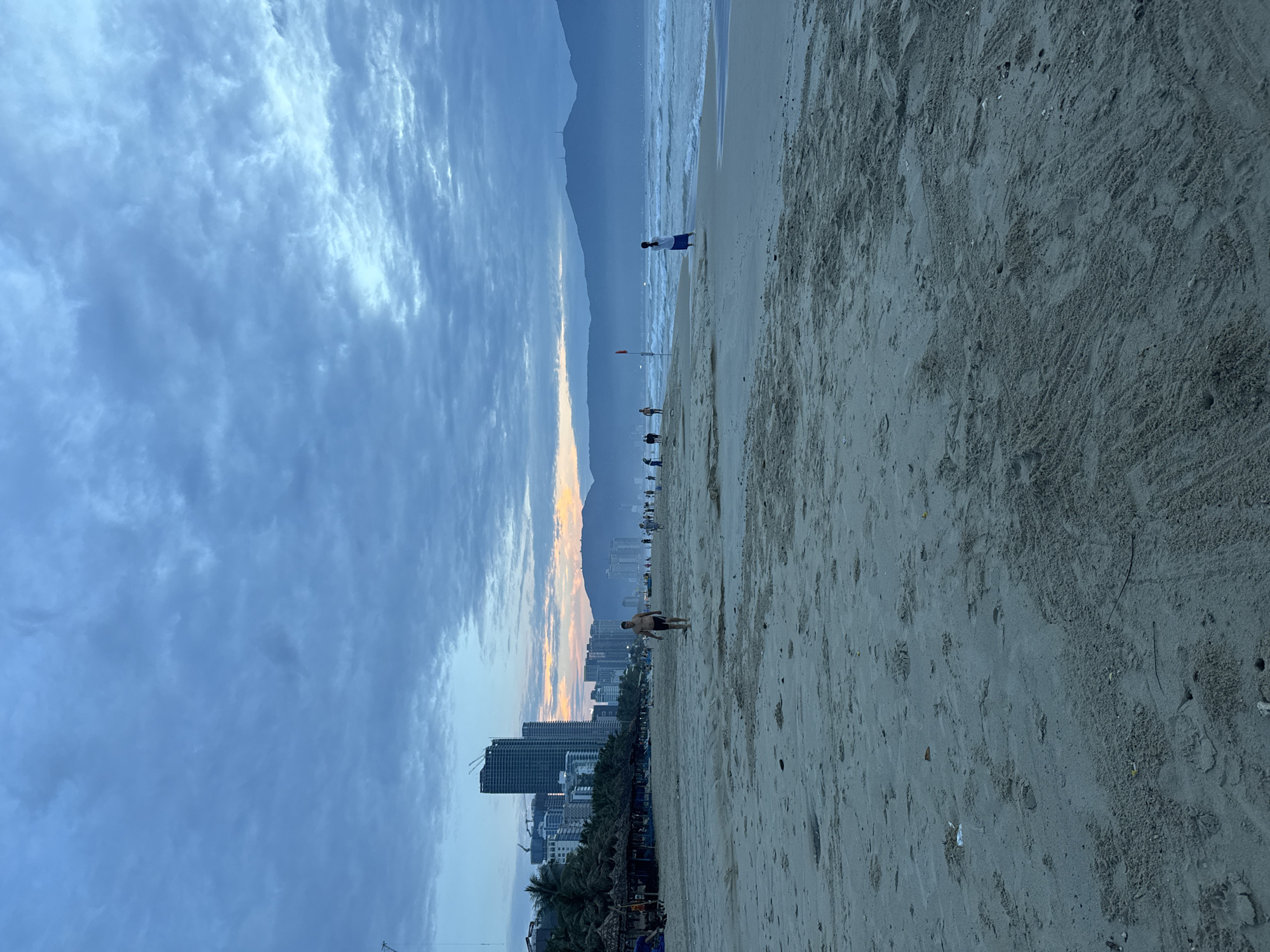 This image depicts a beach scene in Đà Nẵng, Vietnam. The perspective is oriented vertically. The foreground is dominated by sand, exhibiting various patterns and textures. In the middle ground, several individuals are visible along the shoreline, including a person walking near the viewer and another person standing near the ocean. In the distance, the Đà Nẵng cityscape is visible, marked by tall buildings and palm trees, with mountains behind. The sky is cloudy, with hues of blue and orange suggesting sunrise or sunset.