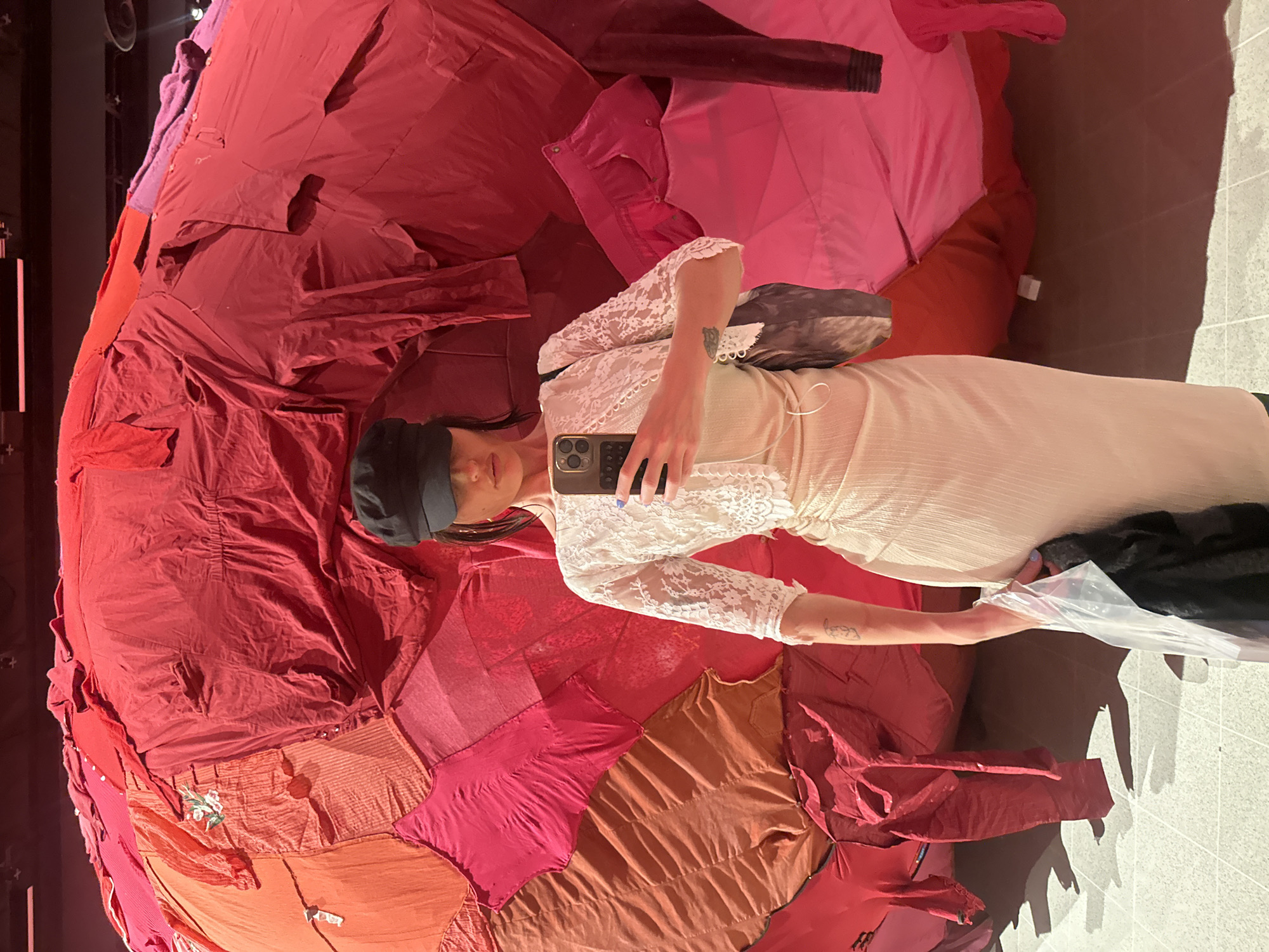 A person in a white dress and a black cap takes a selfie with a smartphone in front of a large, dome-shaped artwork made of various red and pink fabric pieces. The location is in London, United Kingdom. The person is positioned between the artwork and the camera, holding a clear plastic bag in their other hand. The background shows a tiled floor and a dimly lit area.