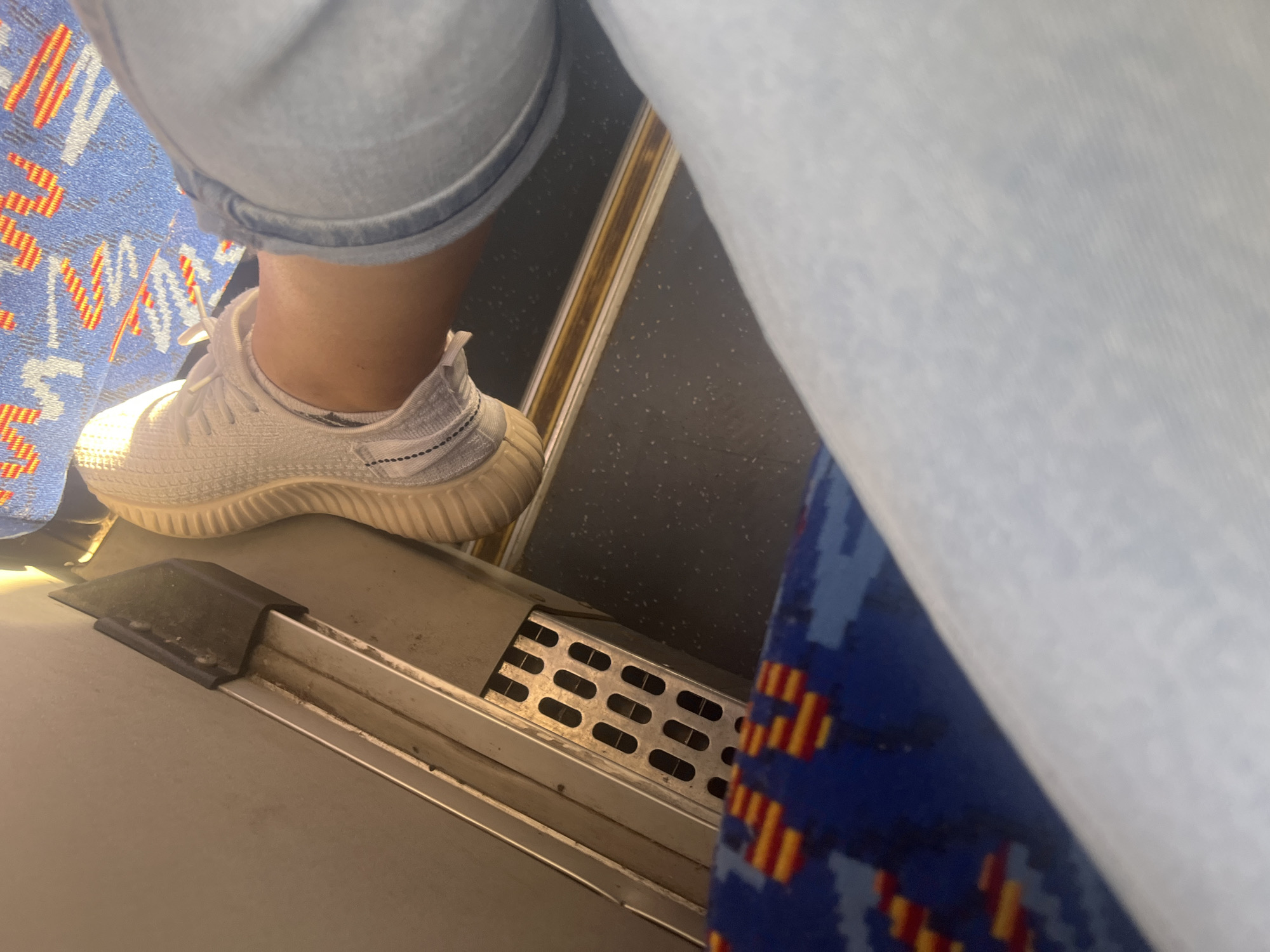The image shows a close-up view of a person's legs and feet inside what appears to be public transportation, possibly a bus or train. The person is wearing white athletic shoes and light blue denim shorts. The floor of the vehicle is visible, featuring a speckled dark grey texture. In the foreground, a portion of a blue seat with a red and yellow pattern is visible. To the left, a metallic structure with small rectangular openings is part of the vehicle's interior, possibly a ventilation grille. The scene is well-lit, suggesting daylight. No specific landmarks or identifying features of Minsk, Belarus are visible in this image.
