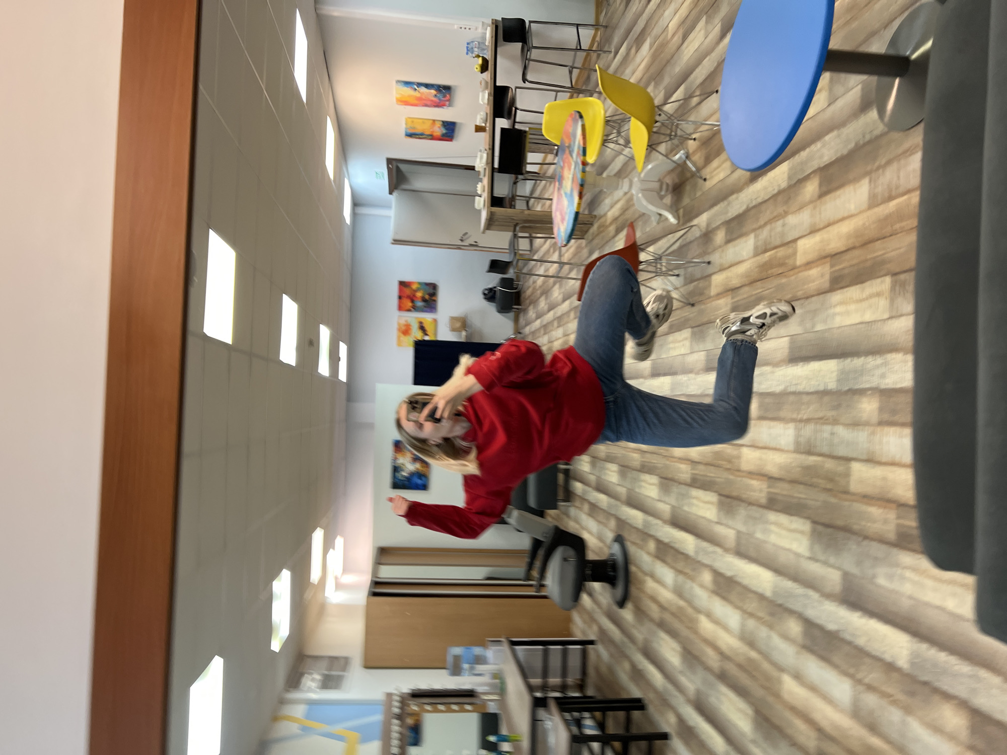 The image, rotated 90 degrees clockwise, captures a modern indoor setting, likely a cafe or common area, located in Khimki, Russia. The flooring consists of light wood-look planks arranged in a horizontal pattern, and the walls are light-colored, featuring several rectangular, glowing light fixtures.
In the mid-ground, a young person with blonde hair is the central focus. They are wearing a red long-sleeved top (sweatshirt or sweater), blue jeans, and light-colored athletic shoes. The person is holding a smartphone to their right ear or near their face, appearing to be talking on the phone or posing for a picture. Their left arm is bent, with the hand in front of them.
The background is furnished with various tables and chairs. There are high-top bar tables with black stools, as well as lower tables including one with a vibrant blue circular top and another with a colorful, abstract patterned surface. A cluster of modern plastic chairs in yellow, white, and red are visible. Several abstract paintings with bright blues, yellows, oranges, and reds adorn the walls. A wooden cabinet or shelving unit and a water cooler are also discernible in the background. In the immediate foreground, part of a grey upholstered chair or couch is visible.
The overall atmosphere is bright and contemporary, suggesting daytime due to the well-lit interior. No legible text is visible within the image.