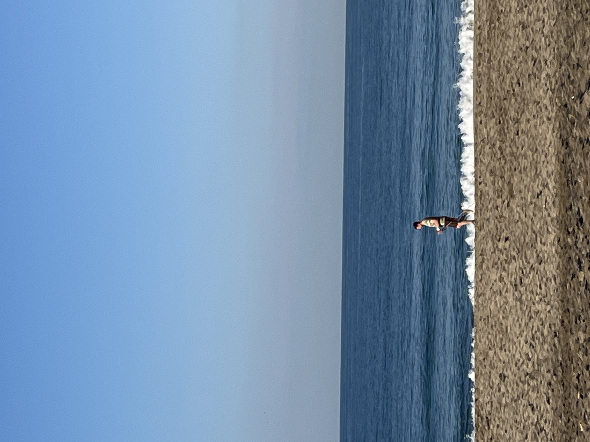 The image shows a beach scene with a man standing at the shoreline. The location is Alanya, Turkey. The sky is clear and blue. The sea is calm with small waves breaking on the shore. The beach is sandy. The man is wearing light-colored clothing. He is standing at the edge of the water, near the point where the waves are breaking.