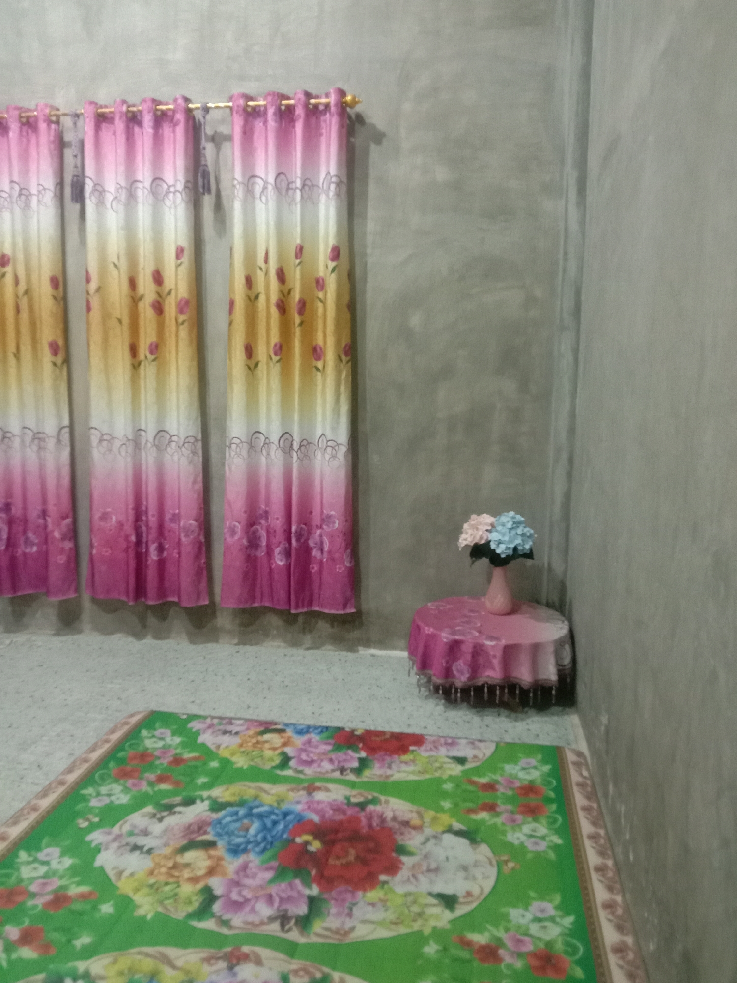 The image shows an interior scene in Lhokjok, Indonesia. The room has gray walls and a tiled floor. Three pink, yellow, and white curtains with floral patterns hang on a rod against one wall. A small, round table covered with a pink tablecloth sits in the corner of the room. A pink vase with a bouquet of blue and pink flowers is placed on the table. A green rug with a floral pattern is on the floor. There are no people visible in the image.