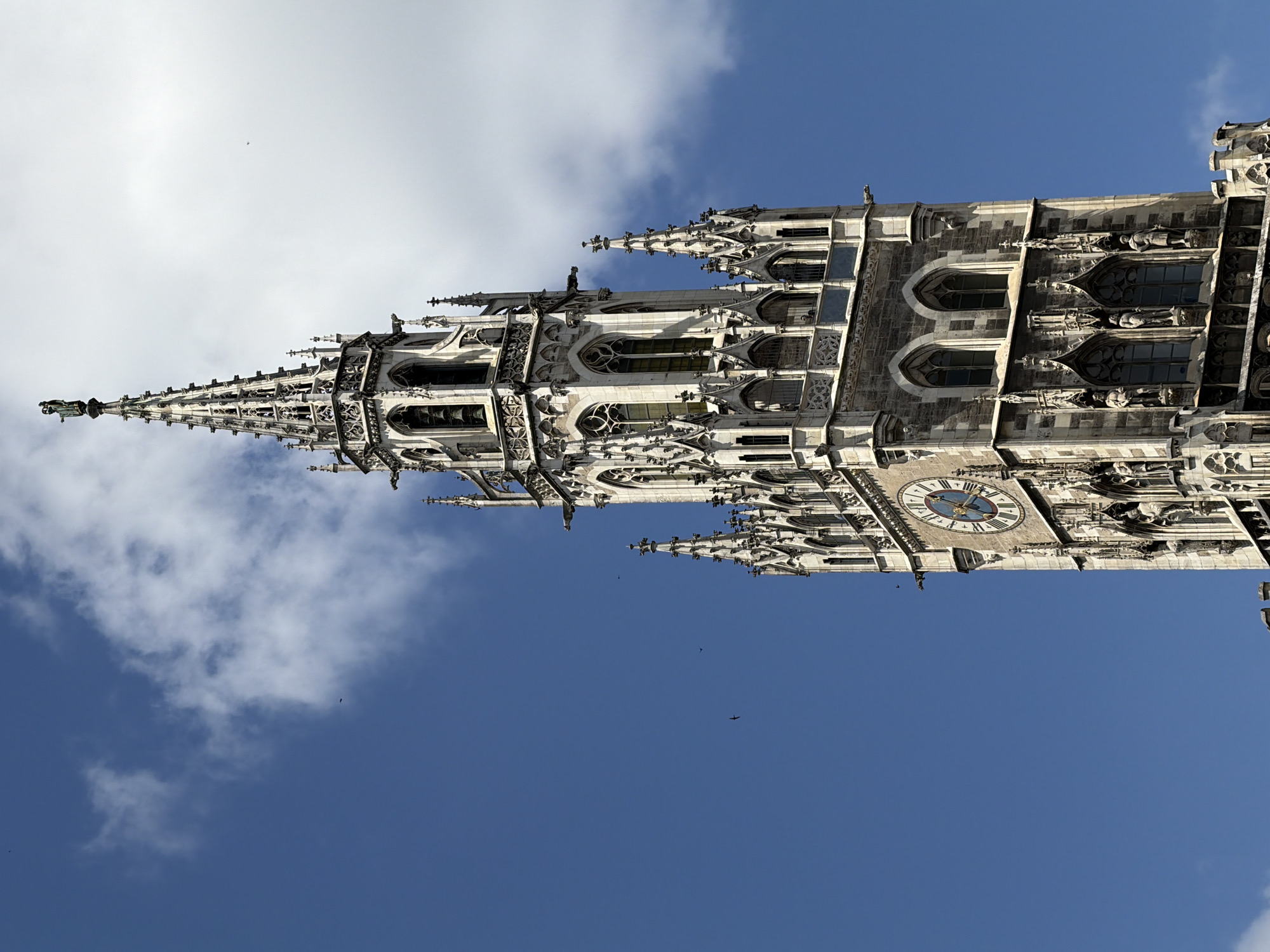 The image is a medium shot of the Neues Rathaus (New Town Hall) in Munich, Germany, viewed from a low angle. The tower of the New Town Hall dominates the right side of the frame, reaching towards a bright blue sky with scattered white clouds. The architecture is Gothic Revival, with intricate stonework, pointed arches, and decorative spires. A large clock face is visible on the tower, with Roman numerals and ornate hands. Small dark figures, possibly birds, are scattered in the sky. The building's facade is made of light-colored stone, with darker stone accents and details. The overall impression is one of grandeur and historical significance.
