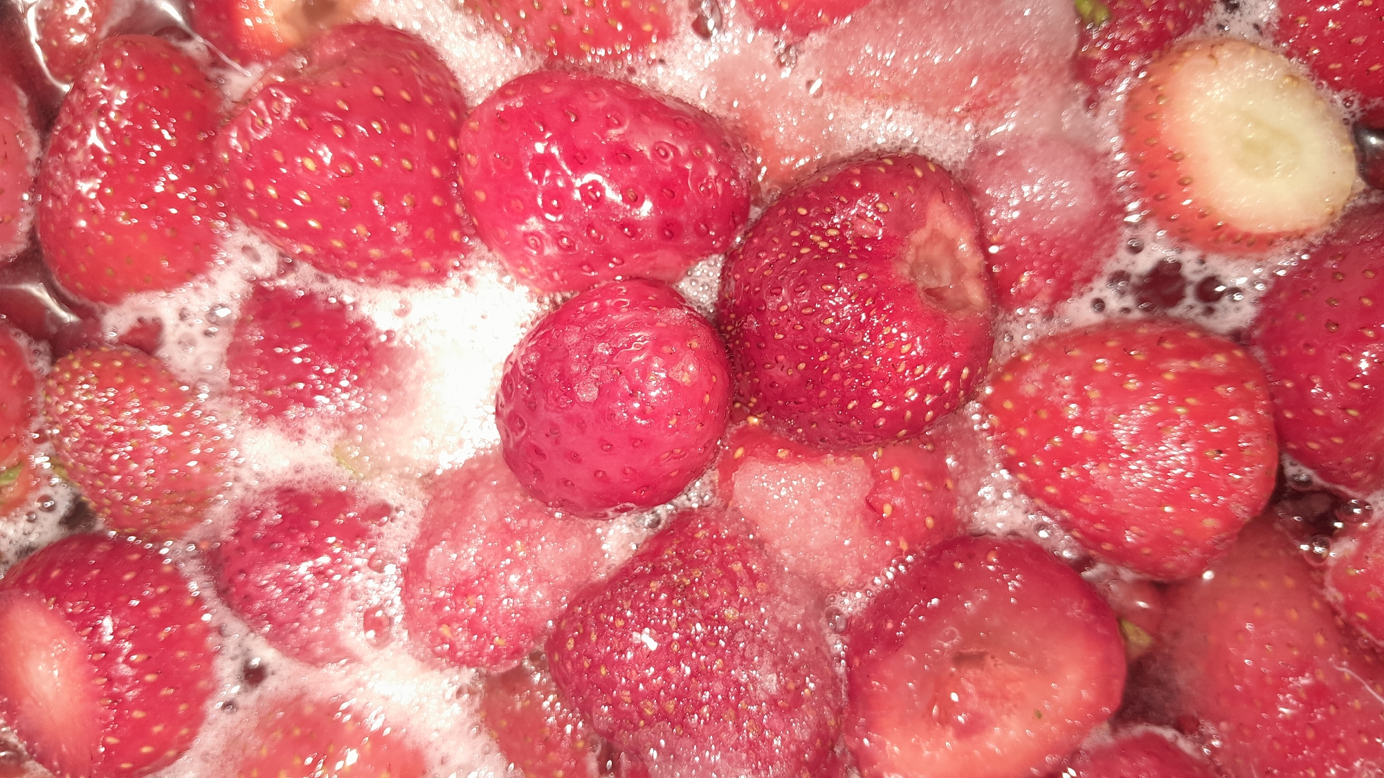 A close-up, overhead shot shows a large quantity of ripe strawberries, many of which are cut in half or quarters, simmering in a bubbly liquid. The strawberries are a vibrant red, with some showing a lighter pink interior. The liquid is frothy and opaque, indicating it's likely a sauce or jam being prepared. The scene appears to be a kitchen setting, possibly on a stovetop, with no specific location in the city of Unknown identified. The focus is entirely on the strawberries and the cooking process.