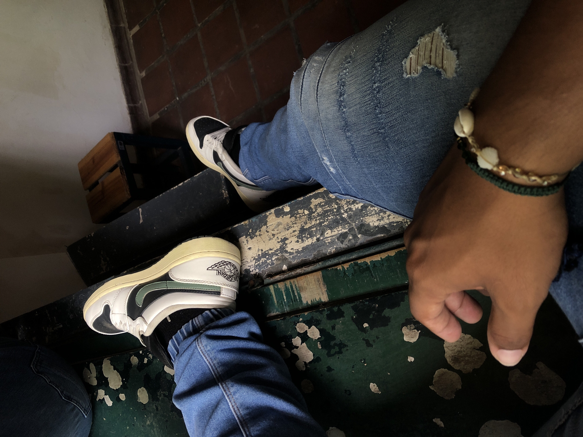 The image presents a low-angle, close-up view of two individuals resting or sitting in what appears to be a worn, rustic setting in El Valle del Espíritu Santo, Venezuela. The primary subject's legs, dressed in distressed blue denim jeans with visible rips on the thigh, are prominent. On their right wrist, a layered bracelet featuring white shells (possibly cowrie), a gold-colored chain, and a dark green braided band is visible.
Both individuals are wearing sneakers. The main subject's shoes are white, black, and dark green with a cream-colored midsole, featuring the distinct "AIR JORDAN" text and wings logo on the ankle collar. A second person's lower leg and foot, also in blue jeans and similar sneakers, are partially visible in the bottom left corner.
They are seated on a dark green surface, likely steps or a bench, which shows significant wear with large patches of paint peeling off. In the background, a rustic wooden crate is visible to the left, positioned against a light-colored wall that transitions into a red brick wall. The lighting is soft and diffused, suggesting a shaded area during the day. The scene conveys a casual and relaxed atmosphere within an aged environment.