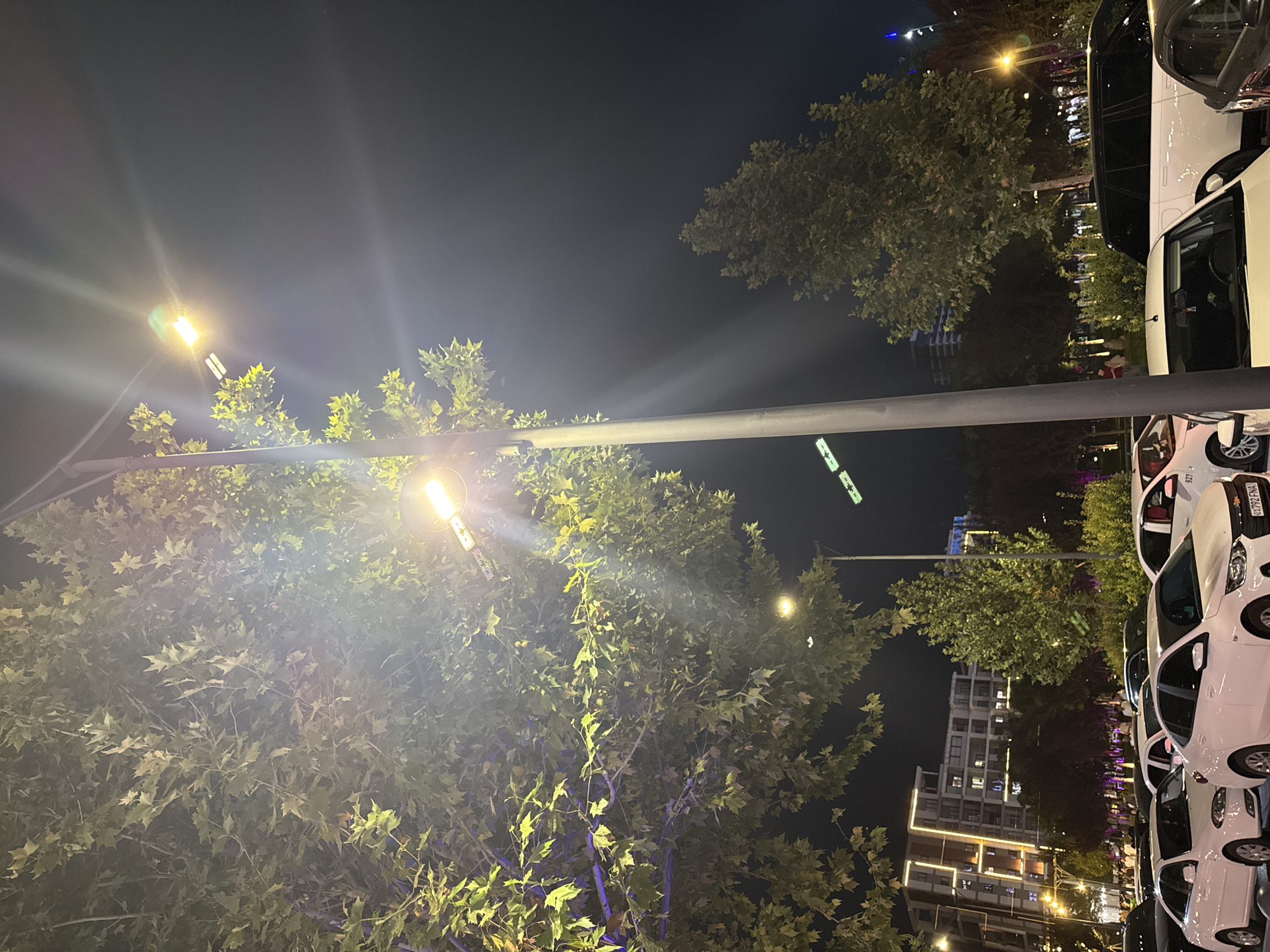 This image captures a street scene at night in Tashkent, Uzbekistan, likely in a modern urban area. A prominent street lamp with bright yellow light illuminates the foreground, casting rays across a large tree with green leaves. In the background, a multi-story building with illuminated windows stands out, suggesting a residential or commercial complex. Several cars are parked along the street, with white sedans and a black SUV clearly visible. The overall atmosphere is one of an active, illuminated urban environment after dark.