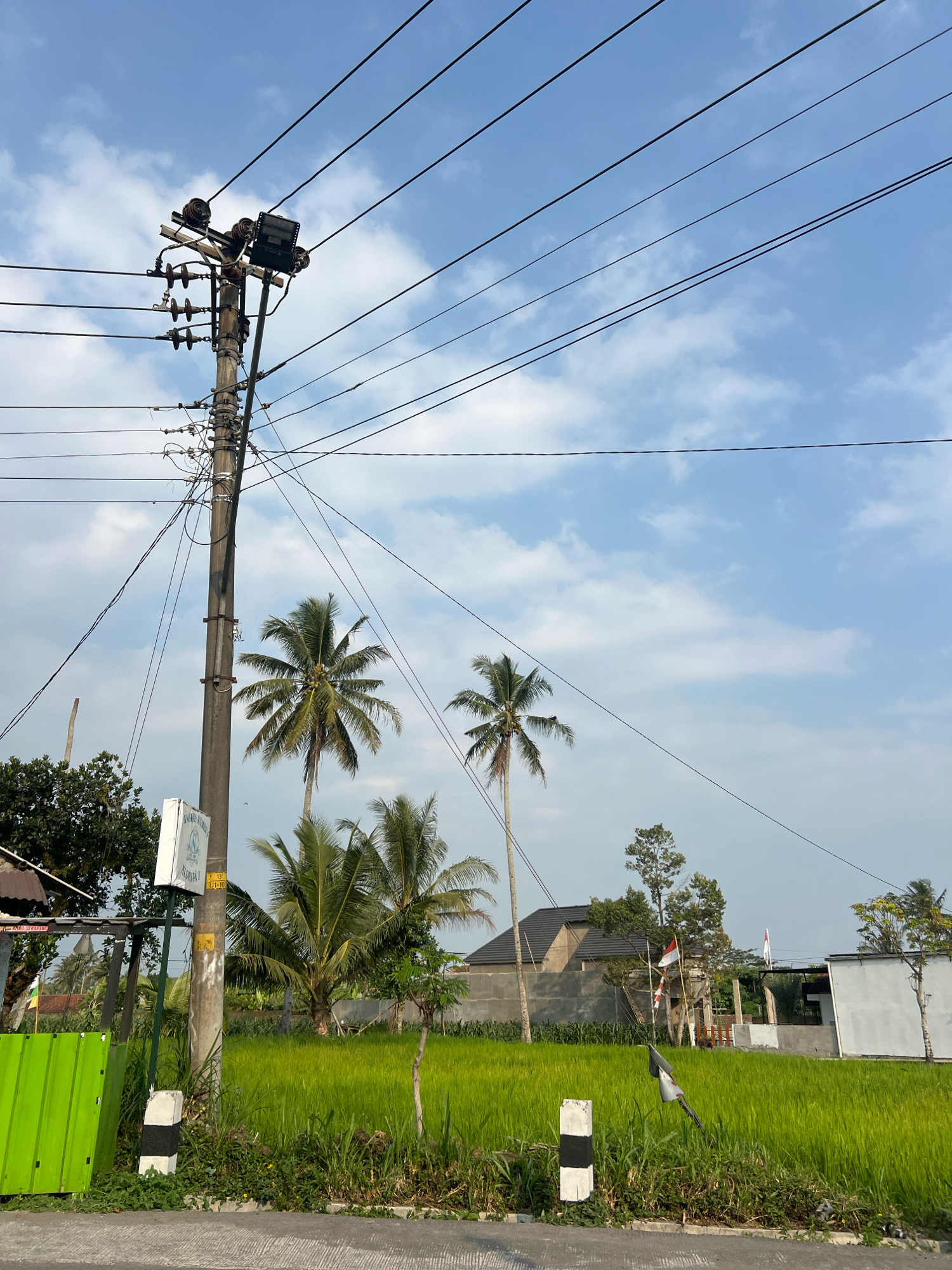 In Wukirsari, Indonesia, this outdoor, eye-level shot captures a bright, slightly hazy daytime scene dominated by a utility pole and its complex network of overhead wires. The pole, made of concrete or a similar material, stands tall on the left side of the frame, supporting numerous insulators and electrical components, including what appears to be a floodlight fixture at the top. Several wires extend from the pole, crisscrossing the sky.
The background features a lush, green rice paddy field, indicating a rural or semi-rural setting. Two mature palm trees are prominently visible within the field, their fronds catching the sunlight. Further back, construction is evident, with partially built structures and concrete walls. Two Indonesian flags, red and white, are displayed on poles, one in front of a building and another further back. The sky is a soft blue, populated by scattered, wispy white clouds. In the foreground, a road or paved surface is visible, with grassy vegetation growing along its edge. Black and white striped markers are placed along the roadside. No people are present in the image.