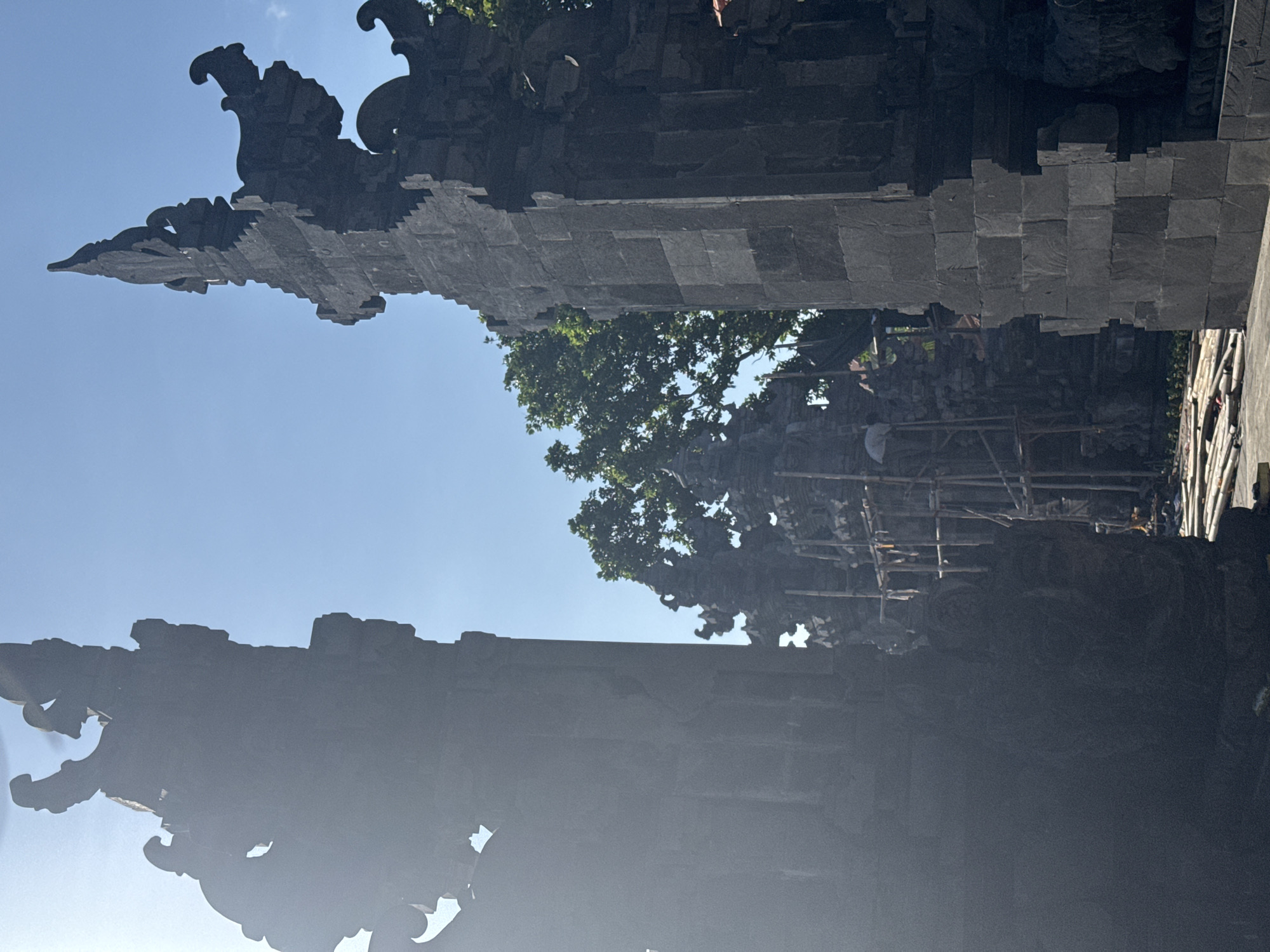 The image captures a scene at a likely temple gate in Kuta Selatan, Indonesia. The composition features two ornate stone structures with intricate carvings, presumably parts of the temple entrance. Scaffolding suggests ongoing maintenance or construction. The structures flank a view of foliage and blue sky. The quality of the image is affected by overexposure and a rotated perspective.
