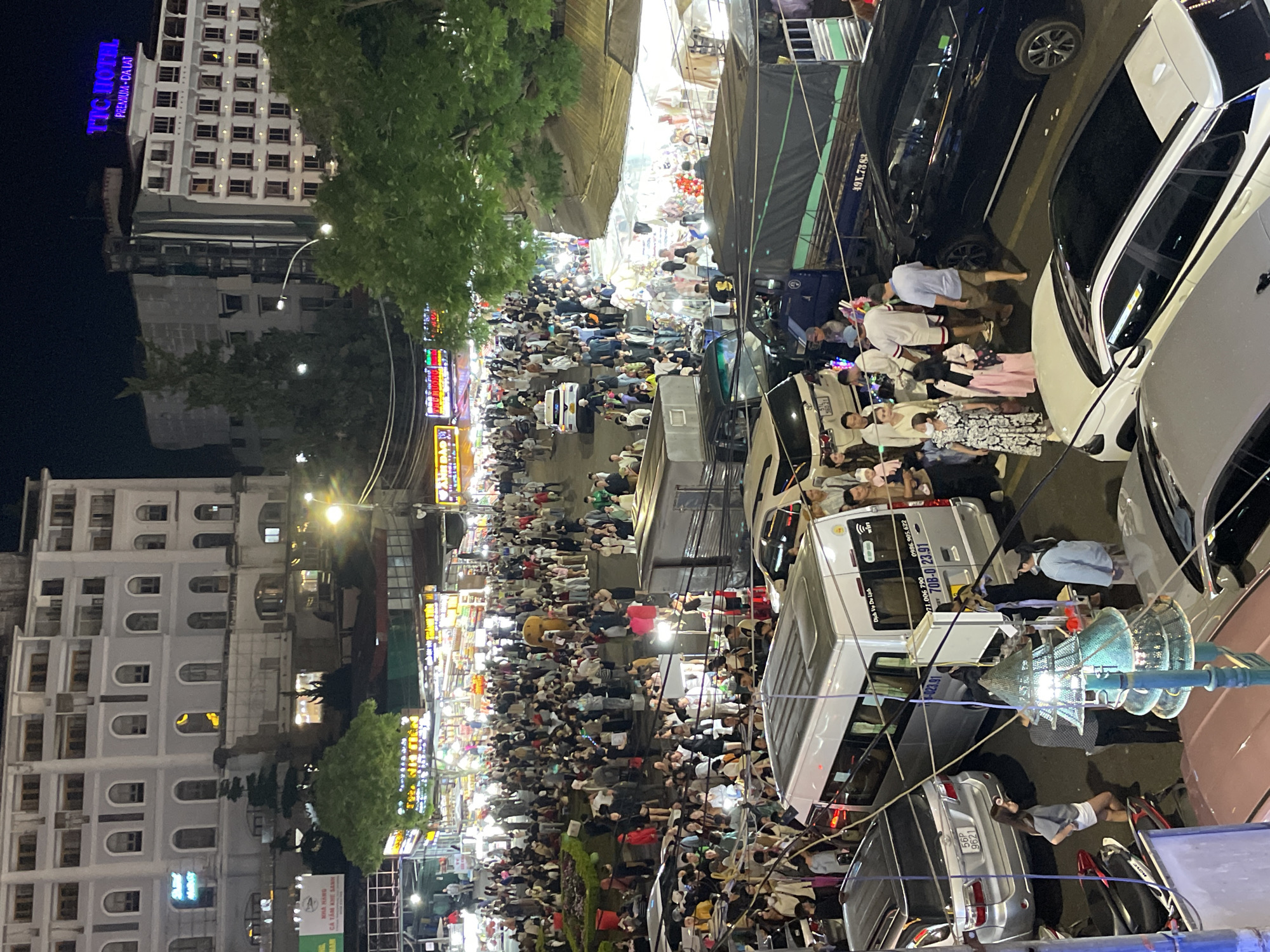This image captures a bustling nighttime street scene in Da Lat, Vietnam, likely a popular tourist area due to the density of people and commercial activity. The foreground features several vehicles, including a silver SUV with license plate "56P 9621," a white van with signage that includes "0986.905.622" and "708-0 23.91," and a dark blue truck with "49X.7383" visible on its side. A prominent object in the lower center is a decorative street light shaped like a Christmas tree.
The street is crowded with a diverse group of people, many appearing to be tourists or shoppers, dressed in casual attire. They are seen walking, standing, and interacting. Some individuals are wearing masks. There are stalls and shops lining the street, illuminated by various lights, indicating a marketplace or night bazaar. The background shows multi-story buildings, including what appears to be a hotel with a sign reading "TTC HOTEL PREMIUM - DA LAT" in bright blue letters. The architecture of the buildings suggests a mix of modern and older styles. The overall atmosphere is lively and vibrant, characteristic of a busy urban night scene. The presence of trees suggests a setting with some greenery, even within the urban environment.