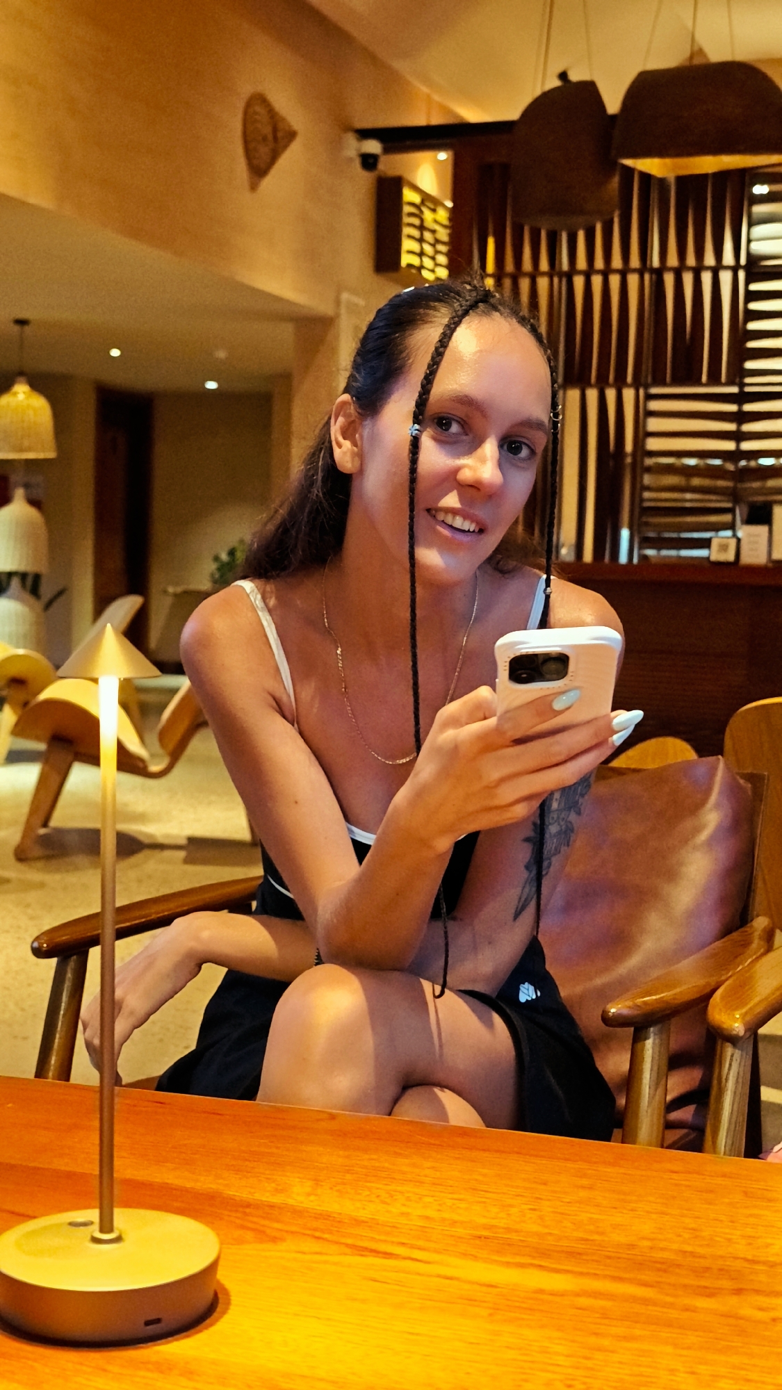 A young woman with dark hair in braids is sitting in a wooden chair at a wooden table, holding a white smartphone. She is wearing a white camisole and a black skirt. The background suggests an indoor setting, possibly a restaurant or hotel lobby, with warm lighting, decorative lamps, and wooden furniture. The environment appears to be upscale and modern. The image does not contain any specific visual cues to confirm its location within Nam Hoa Lư Ward, Vietnam.