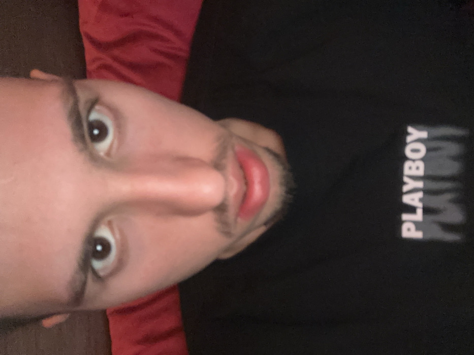 A close-up image captures a young adult male looking directly into the camera. He possesses short dark hair, dark eyes, and a short beard forming a goatee. He is wearing a black crew-neck t-shirt featuring the word "PLAYBOY" printed in light pink or white capital letters across the chest, with a blurred, distorted reflection of the text underneath. The subject's face occupies a significant portion of the frame, indicating a close-range photograph, likely a selfie. His expression is neutral. In the out-of-focus background, a crumpled red textile material is visible behind his right shoulder and head. The scene is located in the city of Varna, Bulgaria.