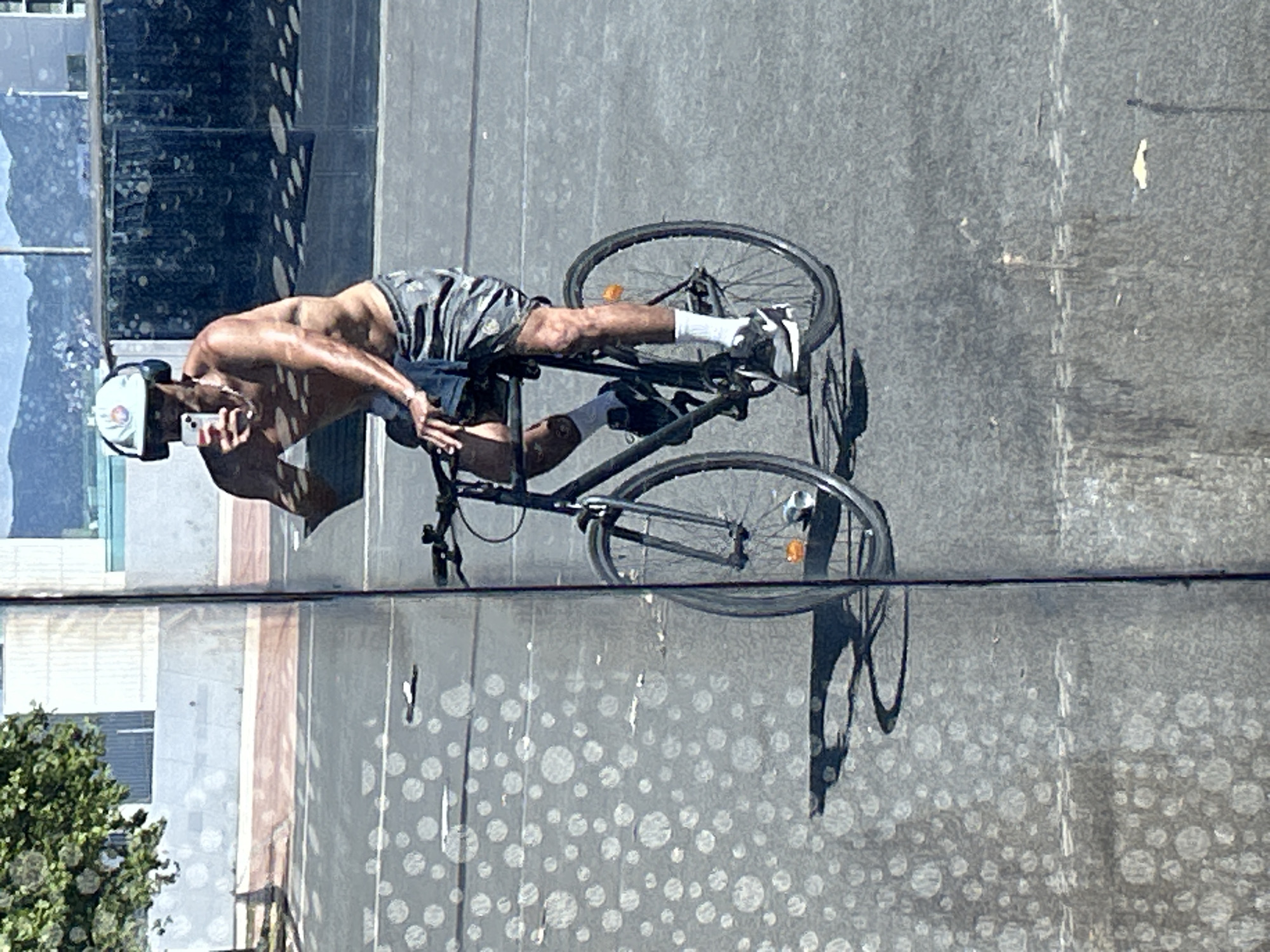 A shirtless person wearing a white baseball cap with a colorful logo, headphones, and patterned shorts rides a black bicycle. They are holding up a white smartphone in their right hand, appearing to take a selfie or record a video. The cyclist is positioned on a paved road, with a shadow cast by the bicycle and rider on the asphalt. In the background, there is a building with reflective windows and trees. The scene appears to be a street in Barcelona, Spain, given the architectural style and natural elements. The reflection in the window shows a blurred cityscape with mountains in the distance. The foreground is textured with circular, light-colored patterns on the asphalt.