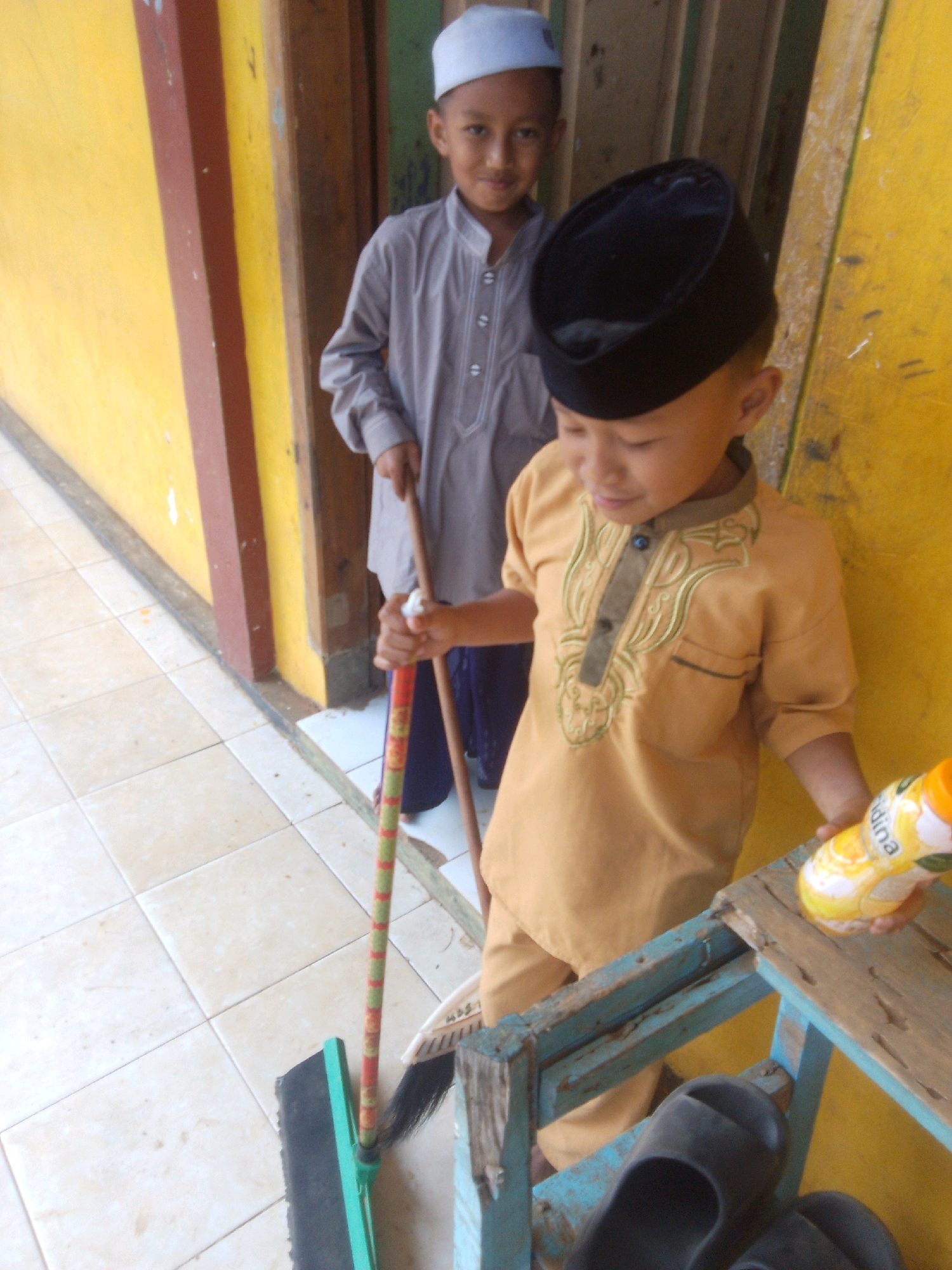 Two young boys are standing near a yellow wall and a wooden door. The boy in the foreground, wearing a tan outfit and a black cap, is holding a bottle of juice and standing next to a broom. The boy in the background, wearing a grey outfit and a white cap, is holding another broom. The scene appears to be in an outdoor or semi-outdoor setting. The floor is tiled. The context suggests this image was taken in Betek, Indonesia, likely in a school or religious institution setting.