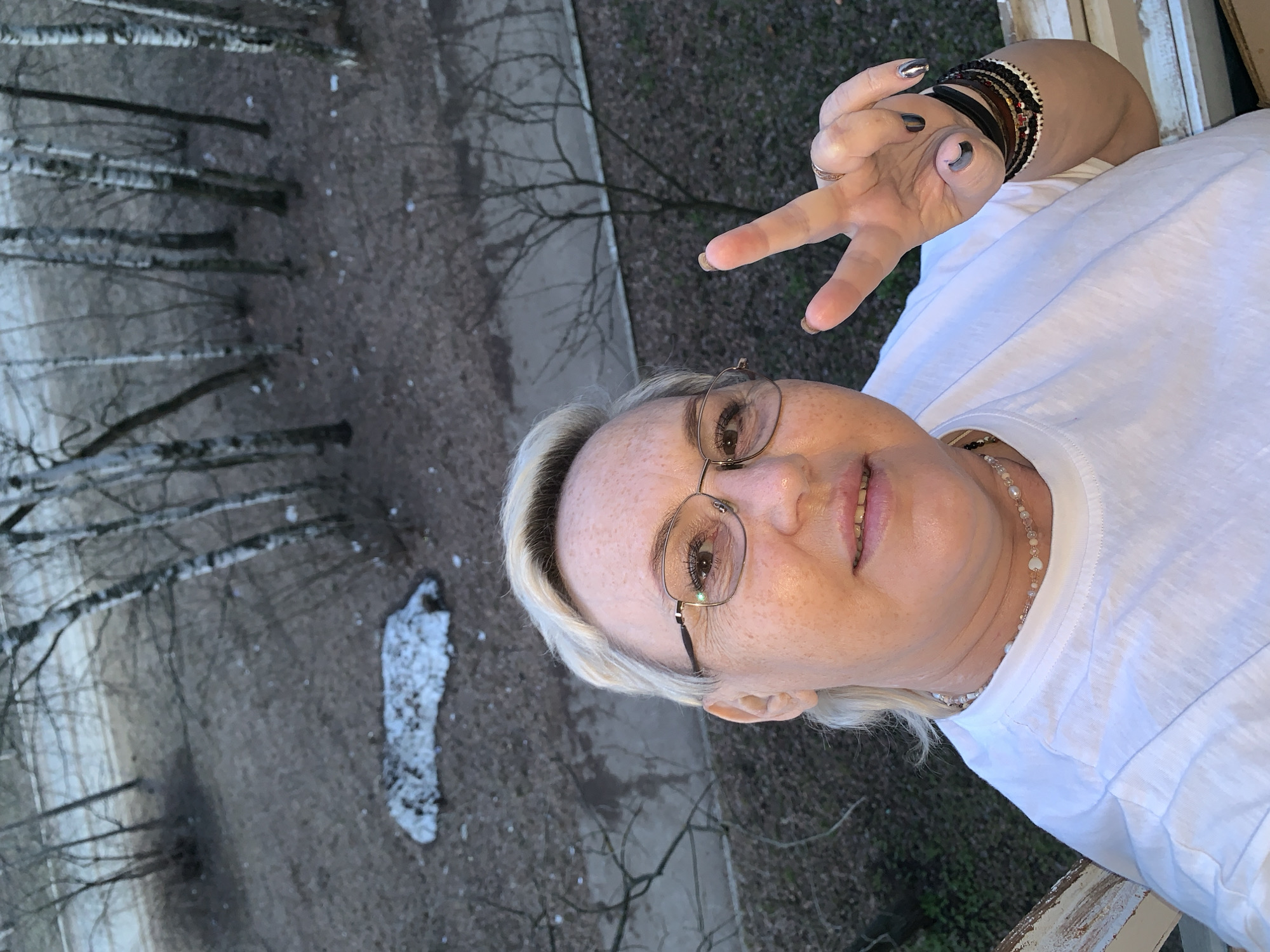 A woman takes a selfie from an elevated vantage point, looking down into a natural landscape. The subject, a woman with blonde hair, fair skin speckled with freckles, and eyeglasses with thin metal frames, smiles at the camera. She is wearing a white t-shirt and a beaded necklace. Her right hand is raised, making a "peace" or "victory" V-sign, with her fingers adorned with dark and metallic nail polish, and her wrist decorated with multiple bracelets, including leather and beaded styles.
The background depicts a winter or early spring scene in what appears to be a park or forested area, consistent with the given location context of Chaikovsky, Russia. Numerous tall, bare deciduous trees, possibly birch, line the view, with their slender branches reaching upwards. The ground is covered in brown earth or fallen leaves, dotted with small patches of white snow or ice. A paved path or road can be seen in the distance. The lighting is natural and diffused, suggesting an overcast day or late afternoon. The woman's smile conveys a positive and cheerful emotion. No visible text appears in the image.