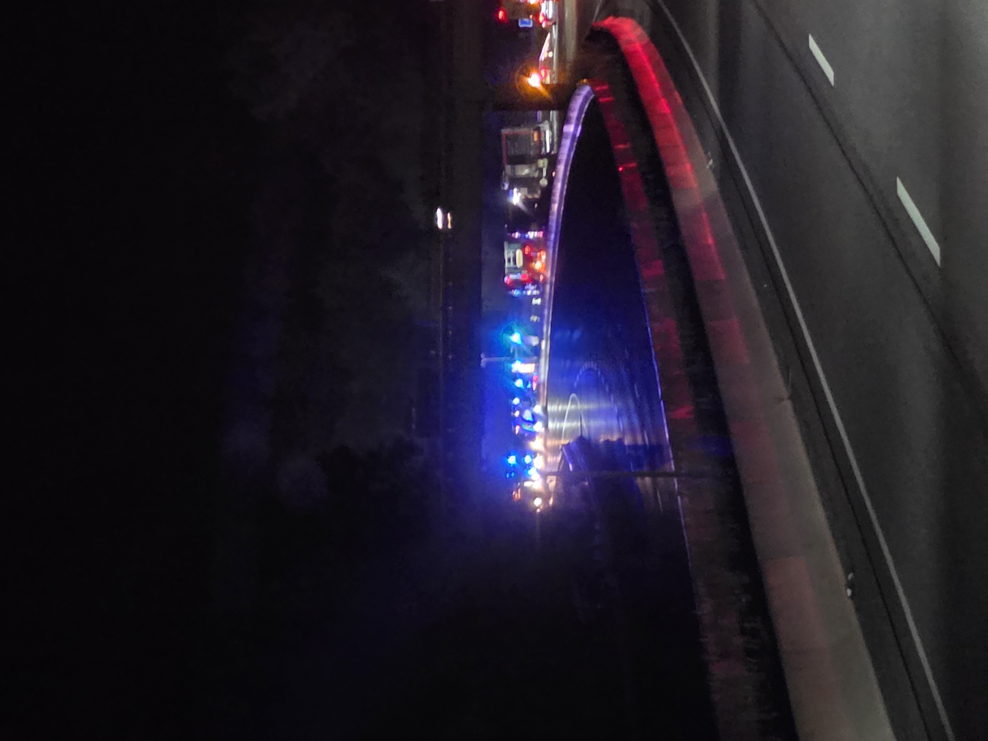In Seligenstadt, Germany, at night, a highway scene is captured. Emergency vehicles, distinguished by their flashing blue and red lights, are positioned along a curved section of the road, possibly indicating an accident or a traffic stop. The vehicles are concentrated under an overpass, with their lights reflecting on the wet pavement. The surrounding environment is dark, with trees visible in the background and the road illuminated by the vehicle lights and streetlights. The overall impression is one of ongoing emergency operations on a thoroughfare.