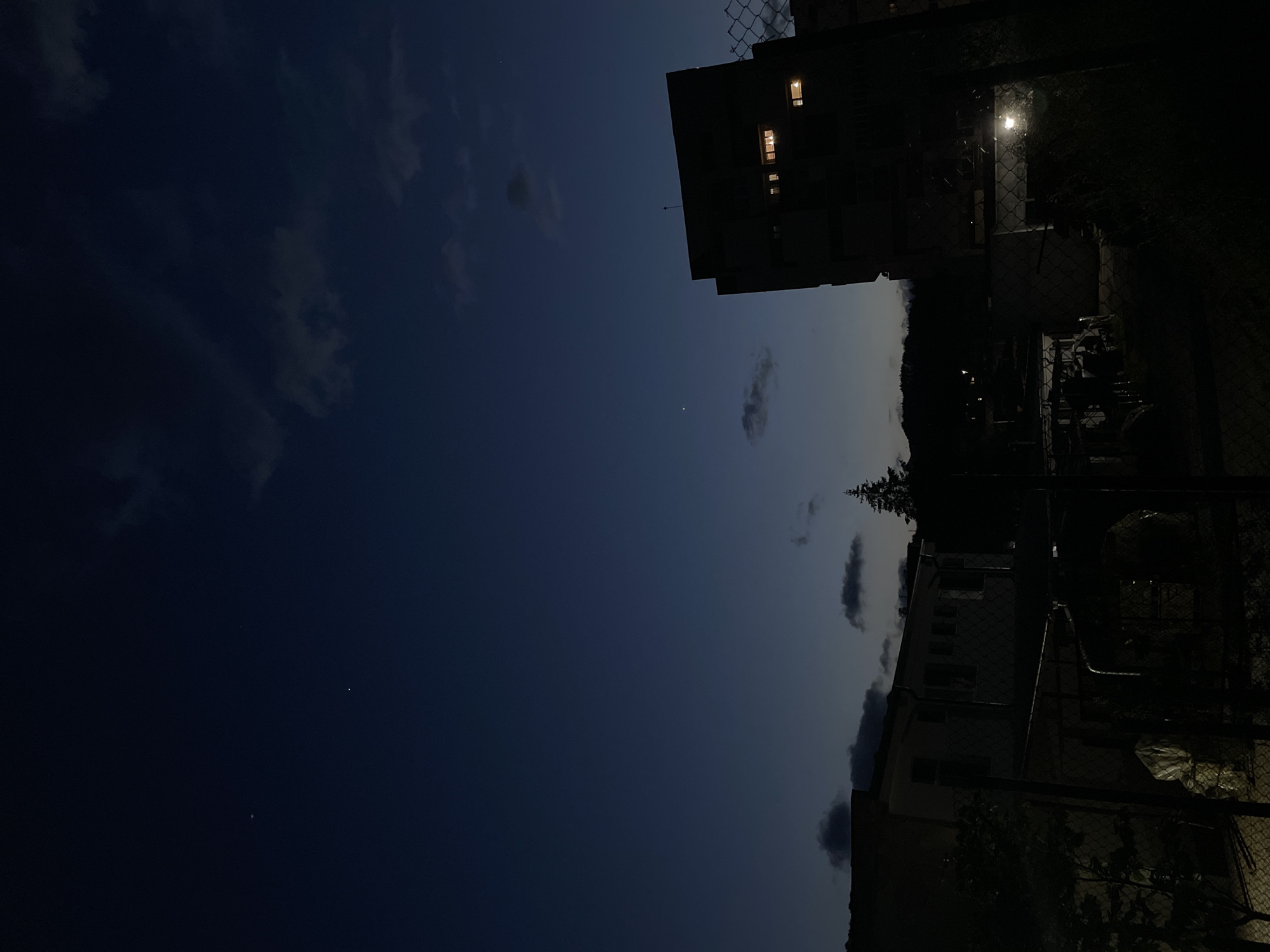 A low-angle, horizontally oriented nighttime photograph shows a view of Zlatograd, Bulgaria. The deep blue sky covers the left side of the frame, with a few sparse clouds. On the right side, several dark buildings and chain-link fences obscure most of the view, with light coming from a lamp post and windows.