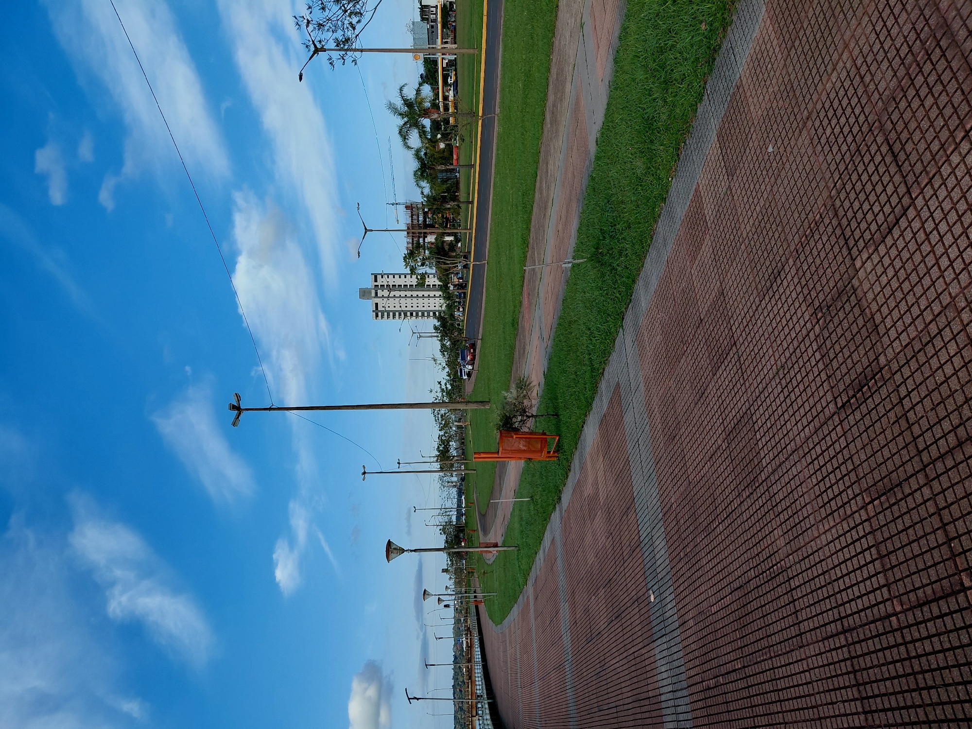 The image depicts a waterfront promenade in Encarnacion, Paraguay, under a bright blue sky with scattered white clouds. The foreground is dominated by a wide, reddish-brown tiled walkway, bordered by a strip of grey concrete. The walkway curves gently to the left, leading the eye towards the distance.
To the right of the walkway is a well-maintained strip of green grass, separated by a low curb. Beyond the grass, a paved road runs parallel to the promenade, with a few cars visible in the distance. A modern, multi-story building stands prominently on the right side of the road, alongside other commercial structures, including what appears to be a Shell gas station. Palm trees and other deciduous trees are interspersed along the roadside, adding to the tropical feel of the environment.
Several lampposts are strategically placed along the promenade and roadside, suggesting the area is well-lit for evening use. An orange public trash can is visible near the grass, close to a small, young tree. The presence of a railing along the left edge of the walkway, partially obscured by the curve, indicates it runs alongside a body of water. The overall scene is one of a clean, urban public space designed for recreation and leisure, likely in a well-developed area of Encarnacion.
