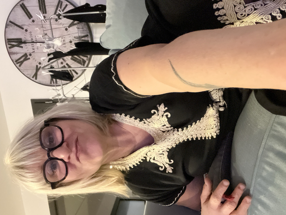 A woman with blonde hair and glasses poses for a selfie. She wears a black top with ornate white embroidery. A large clock hangs on the wall behind her, along with a clothes rack. She appears to be seated on a light blue or green cushion. A tattoo is visible on her arm. The photo is slightly rotated.