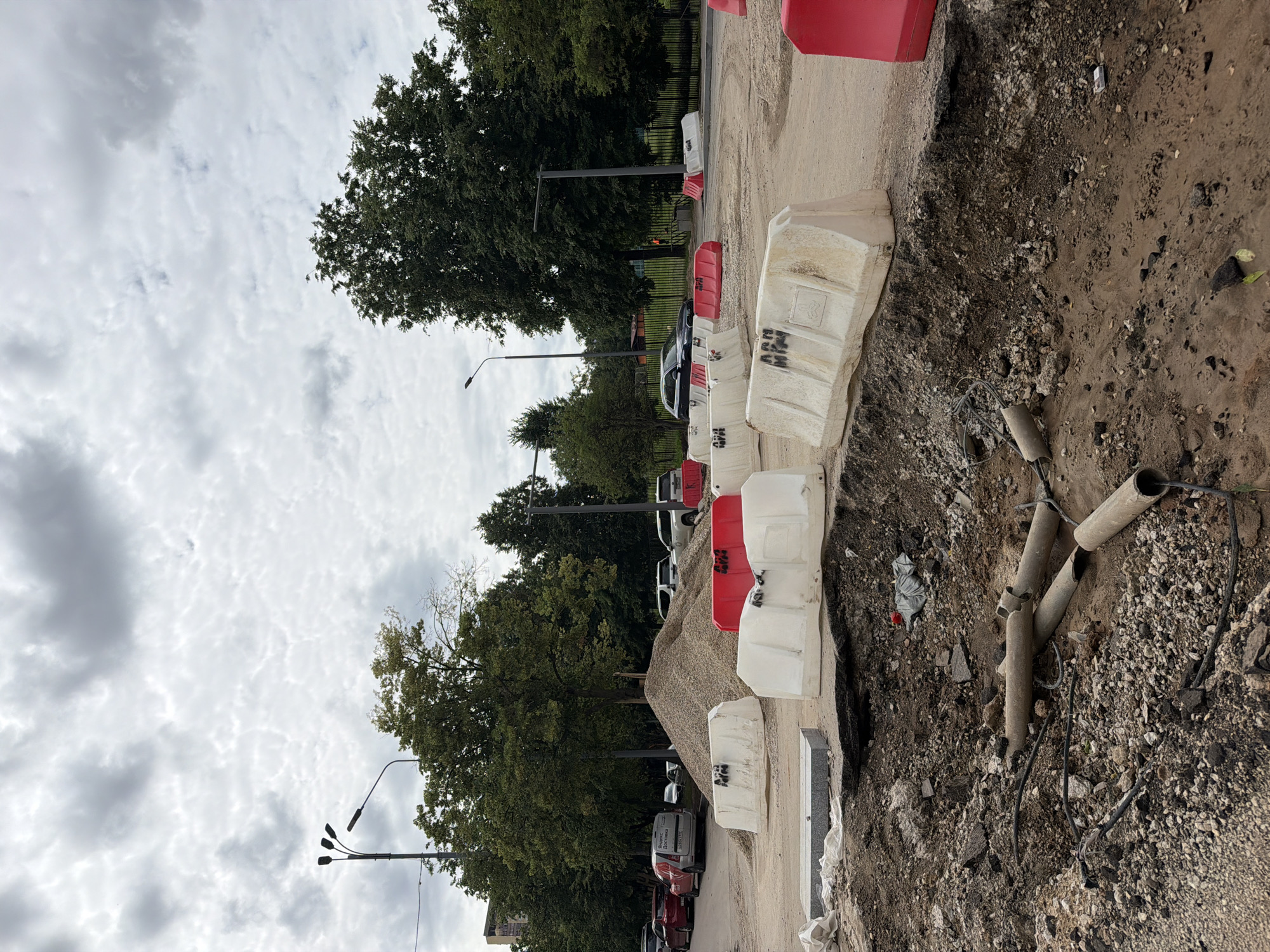 The image depicts a construction site in Moscow, Russia, with a focus on a section of road that has been dug up. Several concrete pipes and electrical wires are visible in the excavated area. The site is cordoned off with a series of white and red plastic barriers. A large pile of gravel sits behind the barriers, suggesting ongoing roadwork. In the background, trees and streetlights line the perimeter of the construction zone. Several vehicles, including cars and a van with "Yandex Delivery" branding, are parked along the side of the road. The sky is overcast with heavy clouds.