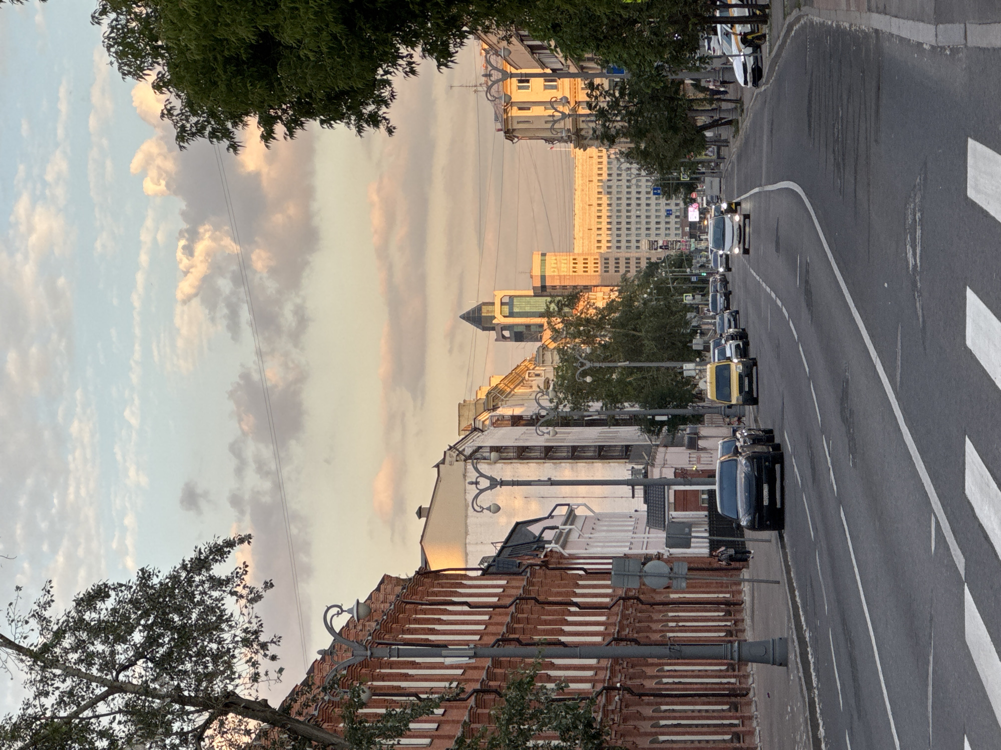 The image captures an urban street scene during the golden hour, with warm, soft light casting long shadows and illuminating buildings. The perspective looks down a street lined with a mix of buildings, ranging from older structures with reddish tiled roofs and ornate streetlights in the foreground to more modern, multi-story buildings and high-rises in the background. Green trees partially frame the view from the top left and bottom right, adding natural elements to the cityscape.
The sky displays a serene blend of light blue, soft pink, and muted gray clouds, reflecting the late afternoon or early evening light. Overhead power lines are visible against the sky.
The asphalt road features a clear white solid line separating traffic lanes and white dashed lines, with a prominent pedestrian crosswalk marked by broad white stripes in the foreground on the right. A variety of vehicles are present: several cars are parked along the side of the road, including a yellow vehicle (possibly a taxi) and a black car. The license plate on the black car reads "o729xx 777," with "777" being a Russian region code for Moscow. Further down the street, a white car with illuminated headlights suggests ongoing traffic. Ornate streetlights are mounted on the buildings and poles along the road.
The scene depicts a typical urban environment, characterized by daily traffic and a blend of historical and contemporary architecture, all bathed in the tranquil, warm light of what appears to be a late afternoon or early evening in Moscow, Russia.