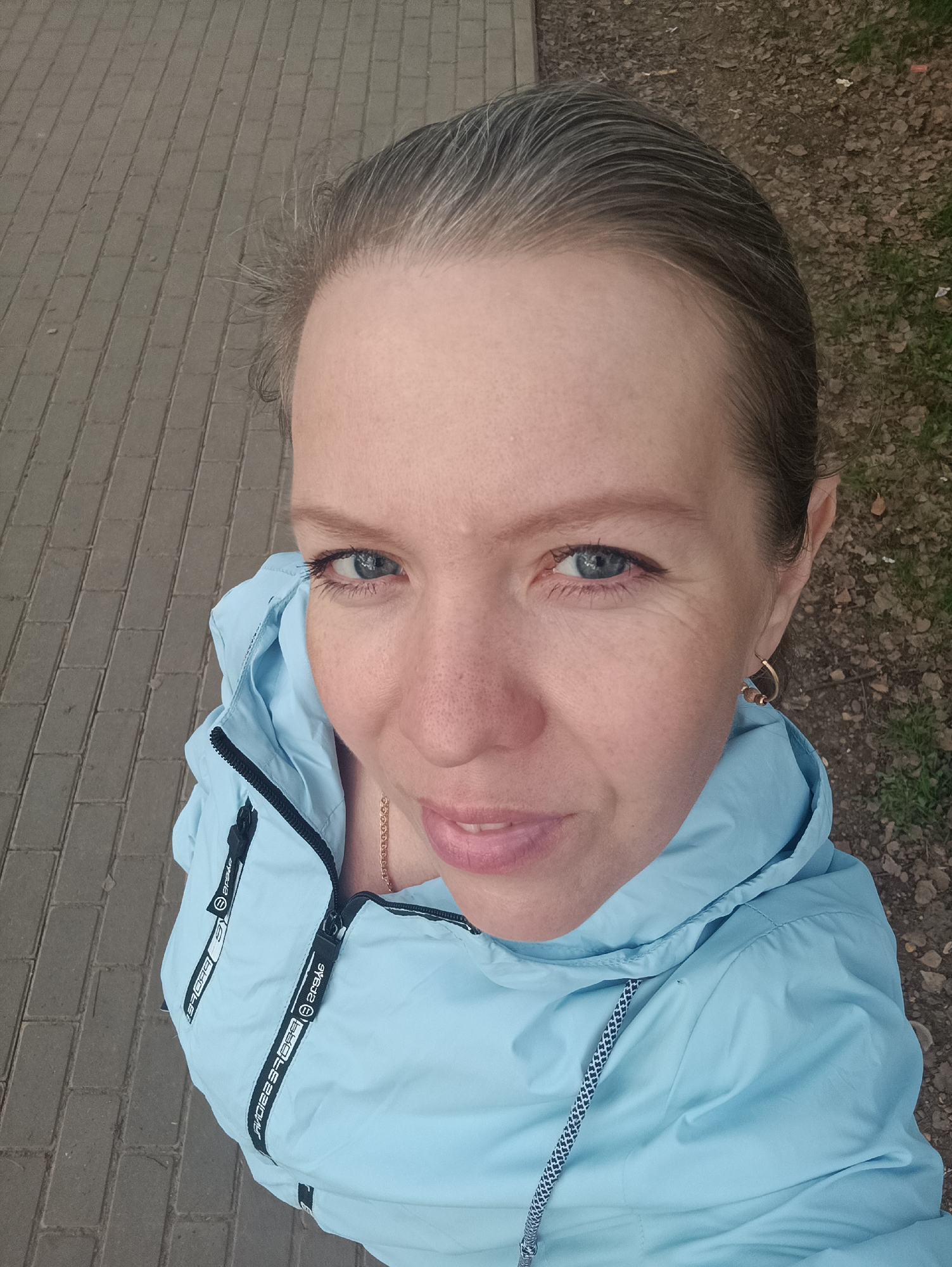 A woman with blue eyes and graying hair is standing outdoors, looking up at the camera. She is wearing a light blue hooded jacket. Part of a brick-paved sidewalk is visible in the lower left of the frame, and a patch of ground covered in leaves is in the upper right. The image was taken in the city of Chaikovsky, Russia.
