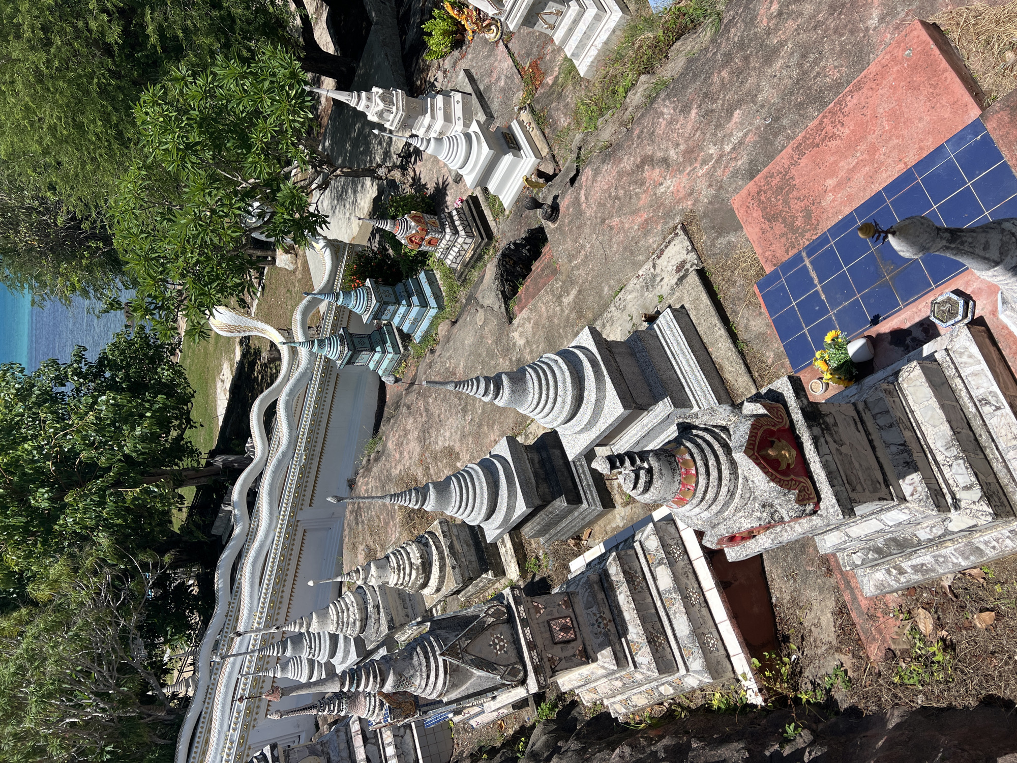 The image displays an elevated view of a Buddhist memorial or temple grounds in Baan Hua Thanon, Thailand. The scene is characterized by numerous small, varied stupa-like structures or chedis arranged across uneven terrain. These structures exhibit diverse designs, materials, and states of preservation; some are plain gray stone, others feature mosaic tiling in shades of white, gray, black, red, and blue, or are painted bright blue and white. Many have tiered bases and conical spires, and some include gold accents.
To the left, a prominent white balustrade, intricately shaped like a multi-headed Naga serpent with gold details, ascends along a visible pathway or staircase. The ground surface comprises concrete pathways, reddish-orange paved sections, and areas of exposed rock and earth with sparse vegetation, dry leaves, and some blue tiled sections.
In the mid-ground and background, dense tropical trees with vibrant green foliage are abundant. Beyond the trees, a clear, turquoise-blue body of water, indicative of the ocean or sea, is visible under bright daylight. A small offering of yellow flowers in a white vase and an incense burner are situated near a stupa in the lower right foreground. No individuals are present in the depicted scene.