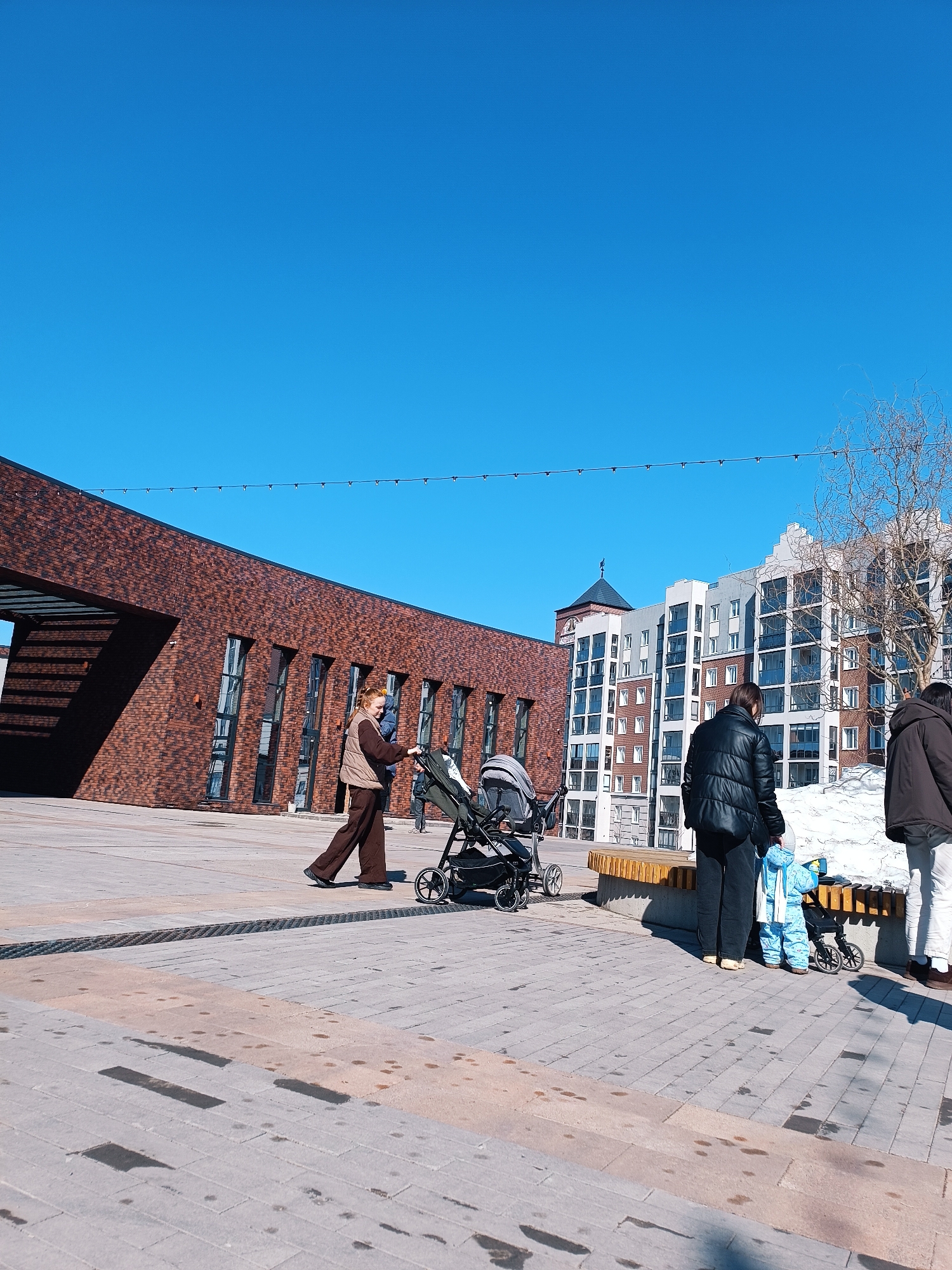 The image depicts an outdoor scene in Gol'yanskiy, Russia on a clear day with a bright blue sky. The location appears to be a public square or plaza, paved with light gray paving stones, some of which have damp spots.
A red-brick building with a distinct architectural style is on the left. It features rectangular window openings and a large, open entrance area. Adjacent to this, a more modern, multi-story building with white, gray, and brick-colored facades is visible.
Several people are present. On the left, a person in a brown outfit is walking with a stroller. To the right of them, another person in a dark coat stands near a wooden bench. A small child in a blue snowsuit is nearby. Further to the right, another individual in a brown coat and white pants is standing.
The interactions between the people are subtle, but they appear to be a mix of passing by, standing, and caring for a child. A patch of melting snow can be seen near the bench. Overhead, strings of small lights are strung across the plaza.