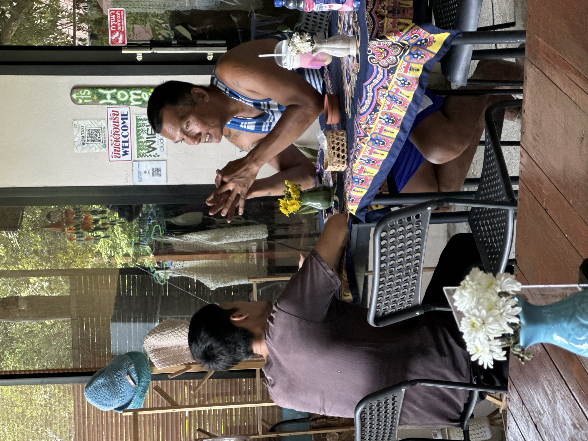 Here is a description of the image:
The image shows a candid scene inside what appears to be a small cafe or restaurant in ตำบลเกาะพะงัน, Thailand. Two people are visible: an older man wearing a blue and white striped tank top and blue shorts sits at a table covered with a colorful patterned cloth. He is leaning forward with his hands together, seemingly in conversation. Opposite him, another individual wearing a brown shirt sits with their back to the camera.
The table has several items on it, including a vase of yellow flowers, a drink with a straw, and other small containers. Behind them is a glass window or door with several signs, including one that says "Welcome" in Thai and English. The OCR also indicates "BOM TAXI" and contact information. Outside the window, one can see greenery and various items for sale, including hats hanging.
The setting appears relaxed and informal, likely a local establishment serving both locals and tourists.