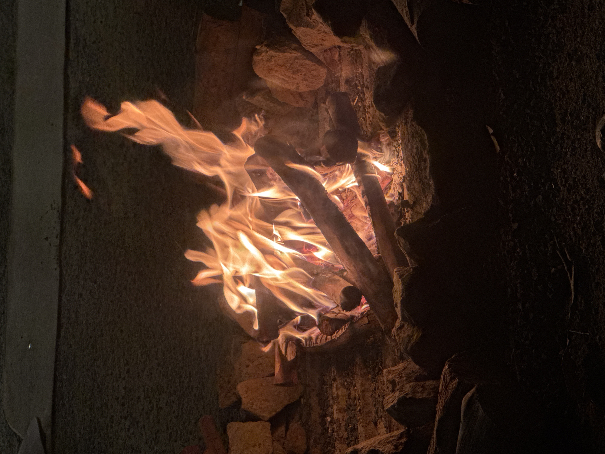 The image depicts a close-up view of a campfire burning in what appears to be an outdoor setting, possibly a fire pit or fireplace constructed from stones. The fire is the central subject, with bright orange and yellow flames leaping upwards, illuminating the surrounding logs and embers. The logs are dry and appear to be burning well. Smoke is visible rising from the fire, mingling with the flames. The background consists of rough, textured surfaces, likely rocks and possibly a dark, gritty wall or ground. The lighting is dominated by the fire's glow, creating a stark contrast between the brightly lit flames and the dark, shadowed areas of the scene. The time of day is clearly night, given the darkness surrounding the fire. There are no people visible in the image. The overall mood conveyed is one of warmth and a primal element of nature. The location context of Nha Trang, Vietnam, suggests this could be a campfire on a beach or in a resort area, though no specific visual cues confirm this. No text is visible.