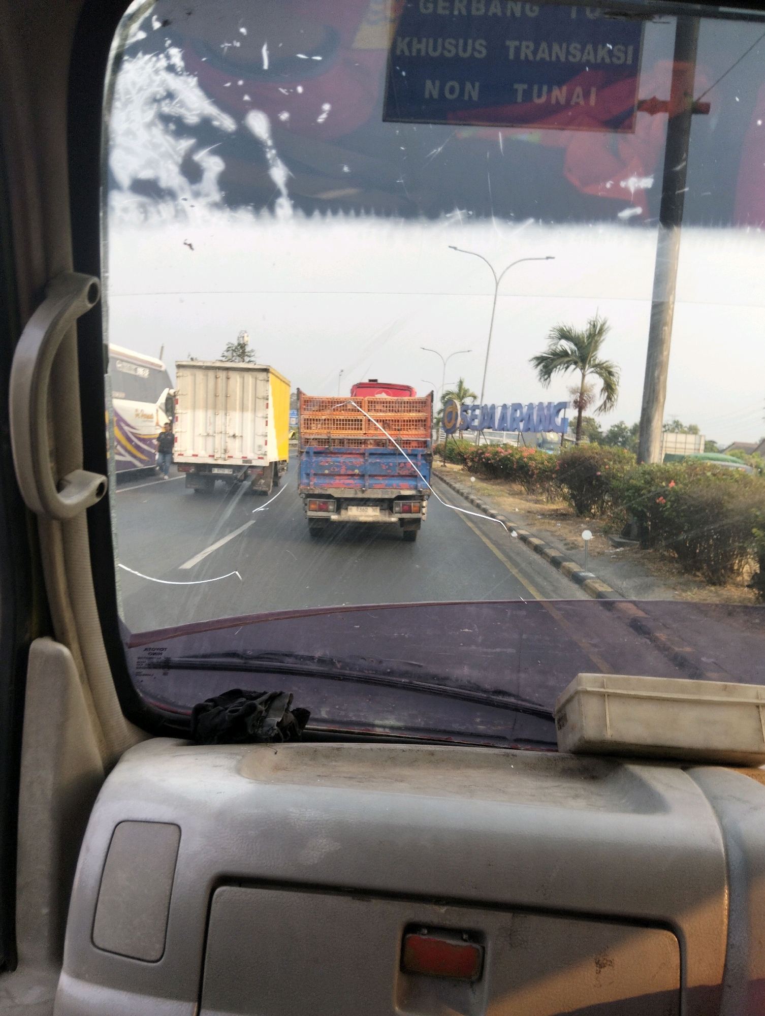 The image is taken from inside a vehicle, looking forward through the windshield, which is dirty and cracked. The perspective suggests a driver's view, with a portion of the vehicle's dashboard visible in the foreground. Outside, on a multi-lane road, several vehicles are present, including a white truck with a yellow container, a blue truck with a red cargo bed, and a bus on the left. A person is seen standing near the white truck. On the right side of the road, a large blue sign with white lettering reads "SEMARANG," indicating the location is the city of Semarang, Indonesia. Behind the sign, there is vegetation and palm trees. Overhead, a pole with streetlights is visible. A dark cloth is placed on the dashboard of the vehicle.