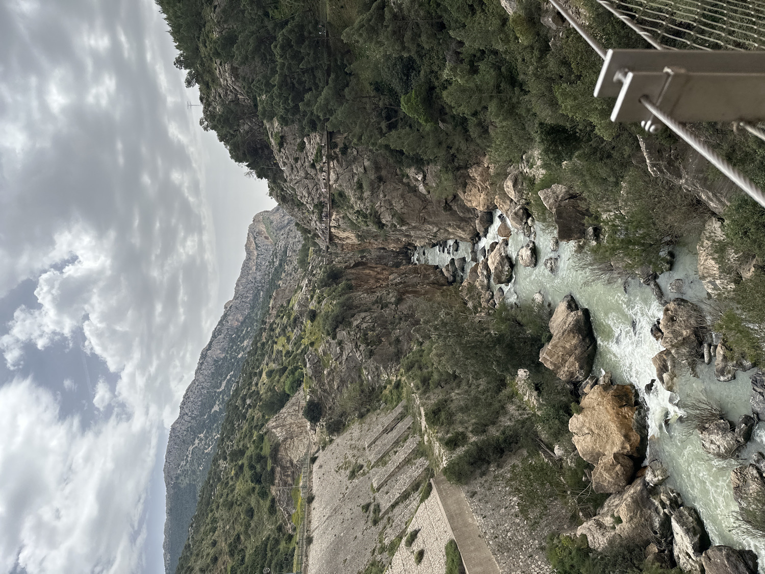 The image presents a dramatic view into a deep, narrow gorge, strongly suggesting the location of El Caminito del Rey near Ardales, Spain. A turbulent river, characterized by its light greenish-blue hue, flows through the bottom of the canyon, churning around numerous large boulders and rocks that punctuate its bed, creating visible white rapids and frothy water.
The steep, rugged walls of the gorge rise sharply on both sides, composed of greyish-brown rock and adorned with patches of dense green vegetation, including trees and shrubs that cling to the cliff faces. High up on the right cliff, a narrow, man-made walkway or path, likely part of the renowned El Caminito del Rey, is visible winding along the rock face, supported by what appear to be metal structures. In the immediate foreground on the right, part of a modern, grey metal railing with wire mesh is visible, indicating the viewpoint from a bridge or viewing platform. Another, older-looking concrete or stone structure runs along the lower left side of the gorge.
The sky is overcast, filled with a mix of light and darker grey clouds, indicating a cloudy daytime setting. No individuals are visible in the scene, which emphasizes the vastness and wild beauty of the natural landscape. There is no visible text within the image.