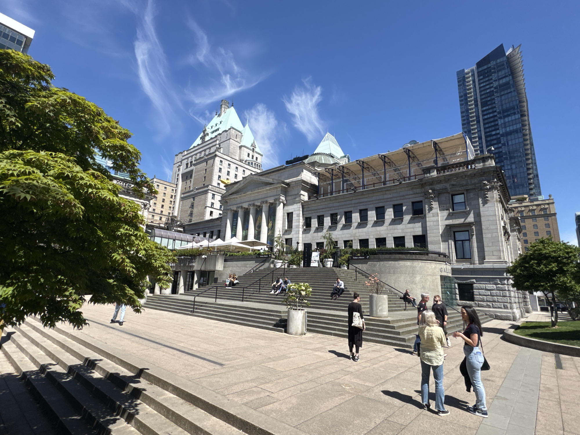 The image shows the Vancouver Art Gallery and the Fairmont Hotel in Vancouver, Canada, on a sunny day with a blue sky and wispy clouds. A modern skyscraper stands in the background to the right of the image.
People are scattered around the multi-tiered steps leading up to the art gallery. Some are seated on the steps, while others stand and chat. The group of three people on the right, standing on the paved ground level, appear to be engaged in conversation. A tree with vibrant green foliage is on the left side of the image, partly obscuring the steps in that area. Another tree is on the right side, at the edge of the paved area.
