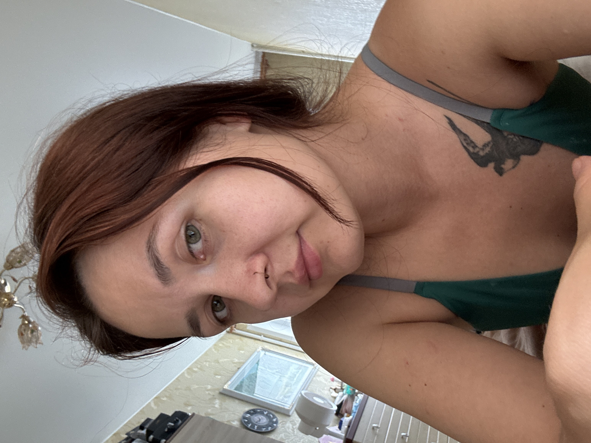 The image shows a close-up, selfie-style portrait of a young woman with auburn hair, green eyes, and fair skin with some blemishes. She is wearing a dark green tank top with gray straps and has a small black tattoo of a bird on her chest. She also has a small nose ring. The woman is looking directly at the camera with a neutral expression.
The background suggests an indoor setting, possibly a bedroom or living area. To the left, a dresser with drawers is visible, topped with various items including what appears to be a small fan and some toiletries. A framed picture hangs on the wall behind the dresser, and a round clock is also mounted on the wall. A ceiling light fixture with decorative elements is visible at the top of the frame. The wall appears to be covered in wallpaper with a textured pattern. The lighting suggests it is daytime, with natural light coming from a window, indicated by the white wall on the right side of the frame.
The image does not provide any specific visual cues that would allow for the precise location within the city of Naberezhnye Chelny, Russia. The interior setting is common and lacks any distinctive landmarks or architectural details that could be used for geolocation.