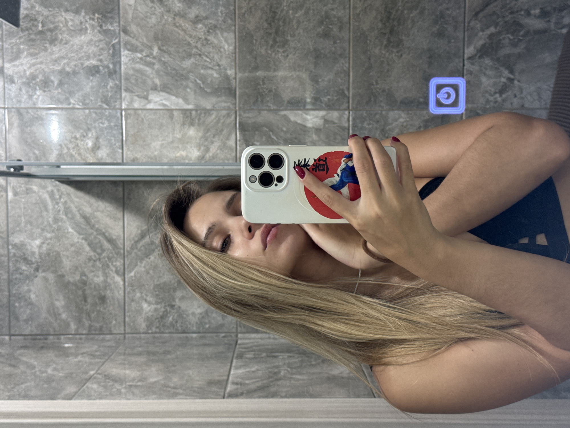 A young woman with long blonde hair takes a selfie in a bathroom with gray tiled walls. She is wearing a black top and has red nail polish. She is holding a white iPhone in her right hand. The phone case has a circular design with an image of a person in a martial arts pose. The bathroom has a mirror with a blue glowing power button icon reflected on it. The location of the image is not specified in the prompt.