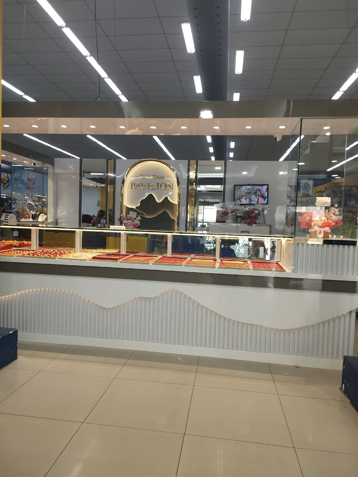 The image shows the interior of a jewelry store, likely a chain called "Pavilion Gold", located in Batam, Indonesia. The store features a well-lit ceiling with fluorescent lights and a clean, modern design. The main display area showcases various gold jewelry items laid out on red and blue velvet trays behind glass counters. Above the counters, a prominent illuminated sign in an arched frame reads "TOKO EMAS PAVILION", with stylized mountain graphics and the text "EMAS" (gold) at the top. There are also floral arrangements, a television screen displaying security camera footage, and a "HALAL" certification sign visible. The floor is tiled with large beige tiles. A portion of a white, decorative wall with vertical ribbed detailing is visible in the foreground, and a dark blue object, possibly a seating or storage box, is present on the left side of the frame. Reflections in the glass indicate the presence of customers and staff within the store.