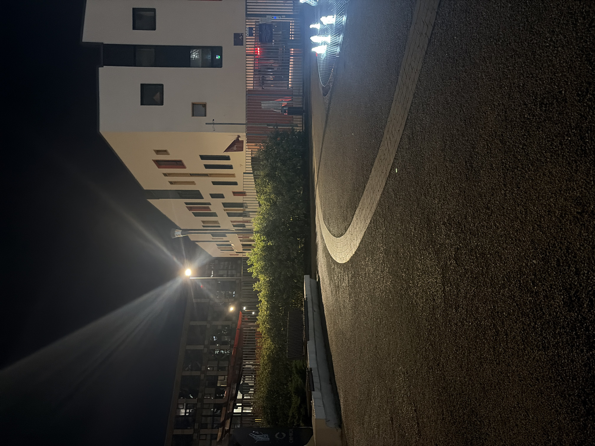 This is a nighttime photograph of an outdoor area with a modern building and surrounding landscaping. A bright streetlamp in the mid-ground casts a strong beam of light upwards, creating visible rays in the dark sky. To the right of the streetlamp is a multi-story building with a white facade and numerous square windows, some of which are lit. In front of this building is a metal fence.
To the left of the streetlamp, another building complex is visible, appearing darker and more industrial with many windows. A curved pathway made of dark asphalt leads from the foreground towards the buildings. Along the left side of the pathway, a long, low concrete planter with a wooden bench is situated amidst dense green bushes. In the far right of the image, a fountain with multiple illuminated jets of water is active. The overall scene suggests an urban or suburban setting at night, possibly a public space or the grounds of an institution. There are no people visible in the image.
