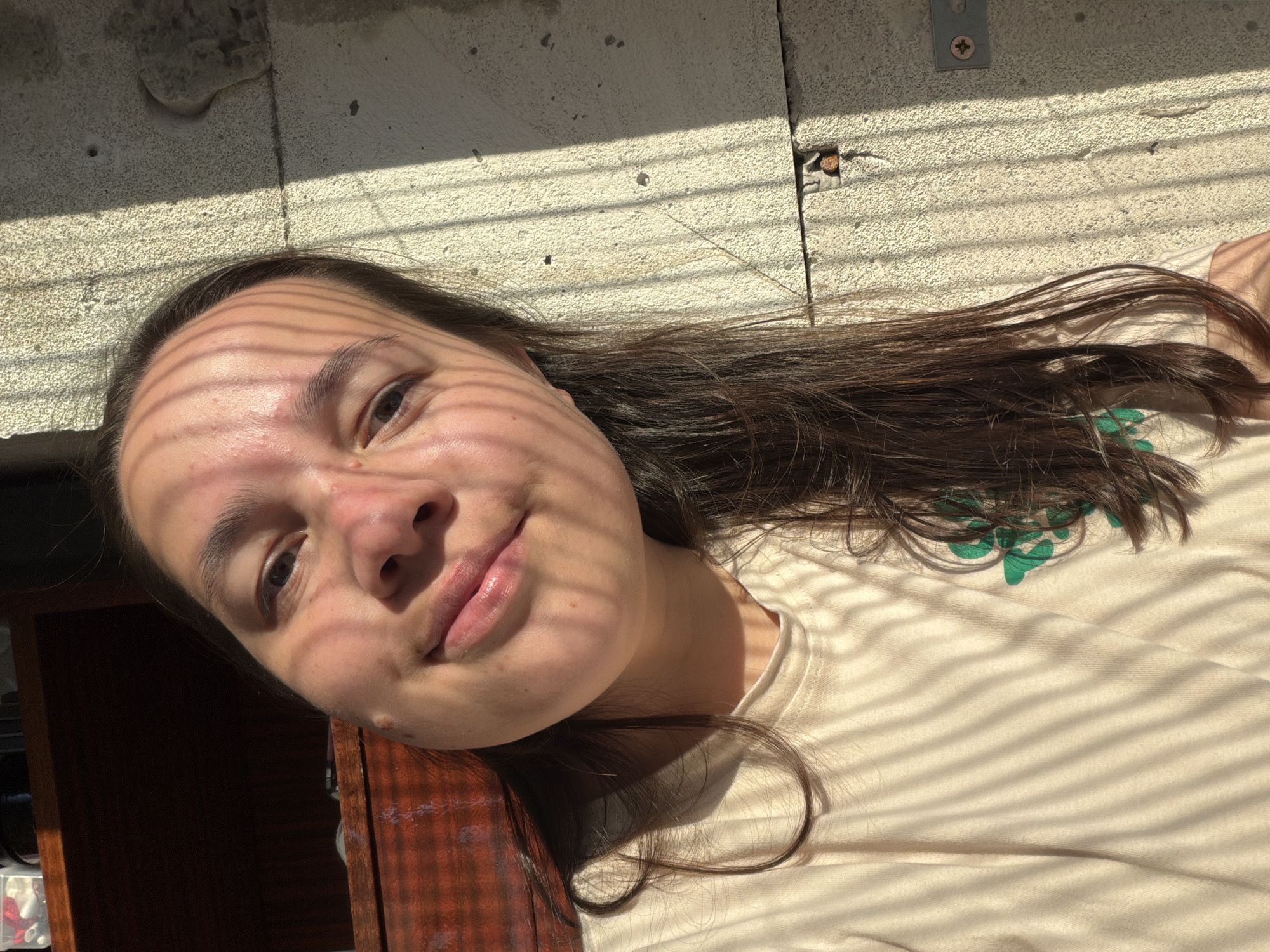 A young woman with long brown hair lies tilted against a wooden surface, likely a piece of furniture. Her face is in sharp focus, illuminated by sunlight filtering through blinds, creating a striped pattern across her face and clothing. She is wearing a light-colored t-shirt with a green design on the shoulder area. The background features a concrete wall and some interior elements, including a dark wooden cabinet. The lighting suggests an indoor setting with strong sunlight entering through a window. There is no information in the image that would indicate it was taken in the city of Chaikovsky, Russia.