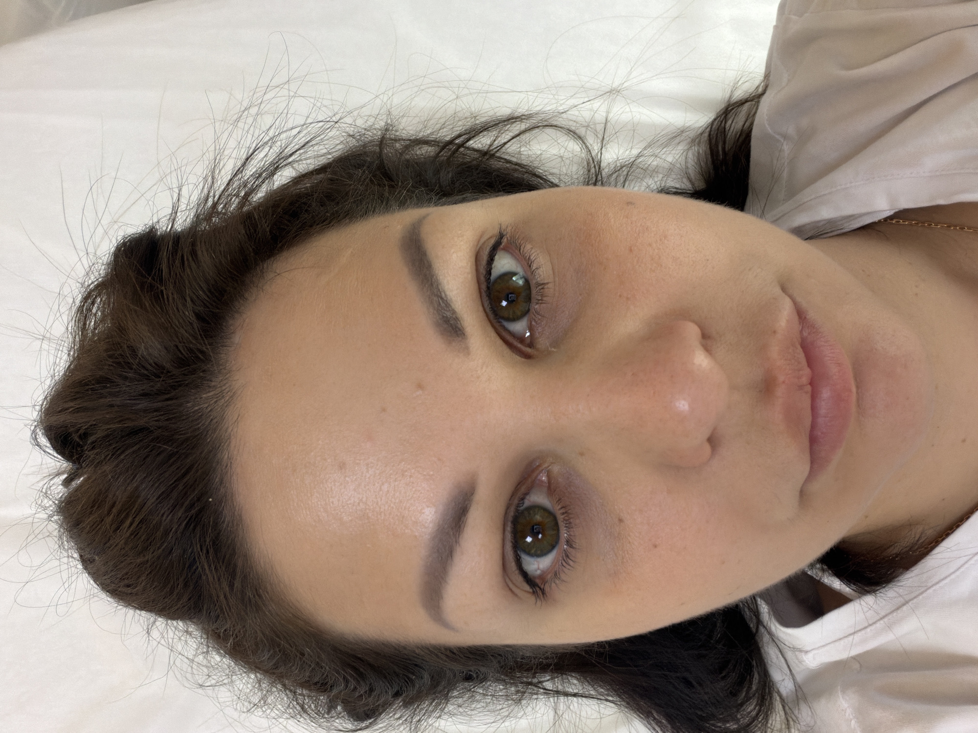 The image displays a close-up, top-down perspective of an adult female's face and upper torso. The individual has dark brown hair, styled back from the forehead, with some flyaway strands. Her eyes are open, featuring brown-hazel irises with visible green/gold flecks and dilated pupils, gazing directly into the camera. Prominent dark circles and slight puffiness are evident under both eyes. Her eyebrows are well-defined and appear cosmetically shaped or filled.
The skin tone is fair to medium, with visible pores and subtle areas of uneven pigmentation or freckles, particularly on the cheeks. A small, light-colored bump is present on the forehead, near the hairline on the left side of the frame. Minimal makeup is visible, including a thin line of dark eyeliner on the upper lash line and mascara on the eyelashes. Her lips are full, naturally pinkish-brown, and slightly parted. The bridge of her nose shows some natural skin sheen.
She is wearing a white collared shirt or blouse, the fabric of which is visible around her neck and shoulders. A thin gold chain with a small, indistinct pendant rests on her shirt below her neck. The background is uniformly plain white, suggesting a soft, featureless surface like a bedsheet or pillow, reinforcing the overhead photographic angle. There are no discernible objects or environmental features within the frame to indicate a specific geographic location, including the city of Sochi, Russia.