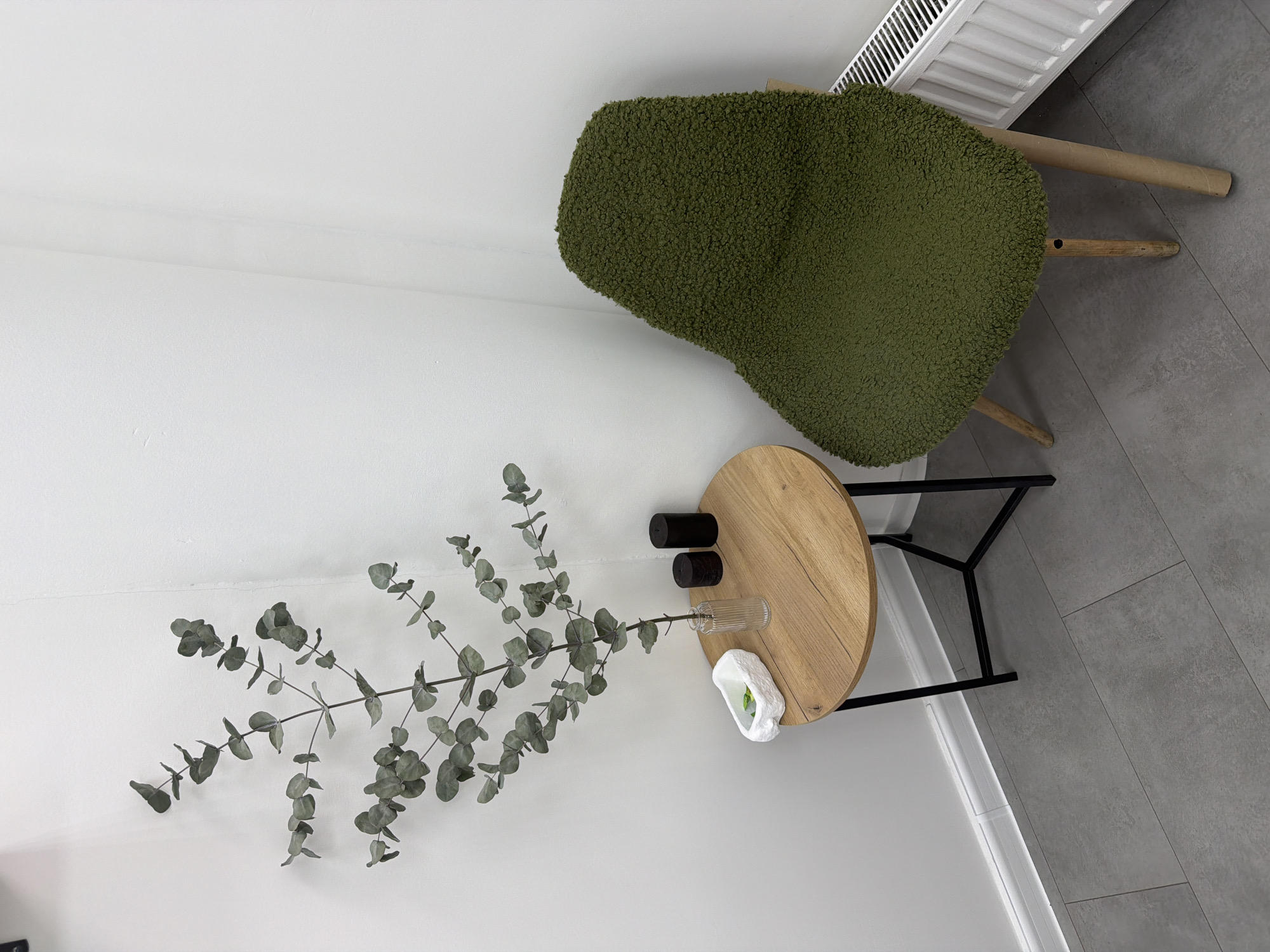 A top-down perspective shows a corner of a room with a chair, a table, and some decorative items. The room has white walls and gray tiled floors. A green, textured chair with wooden legs is positioned in the upper right corner, near a white radiator. A round, light-brown wooden table with black metal legs stands beneath the chair. On the table are two small, dark brown cylindrical objects, a glass vase, and a white, sculptural object, possibly holding a plant. A branch of eucalyptus with silvery-green leaves extends from the table towards the lower left corner of the image, taking up a significant portion of the left side. There is no evidence to identify the location as Moscow, Russia.