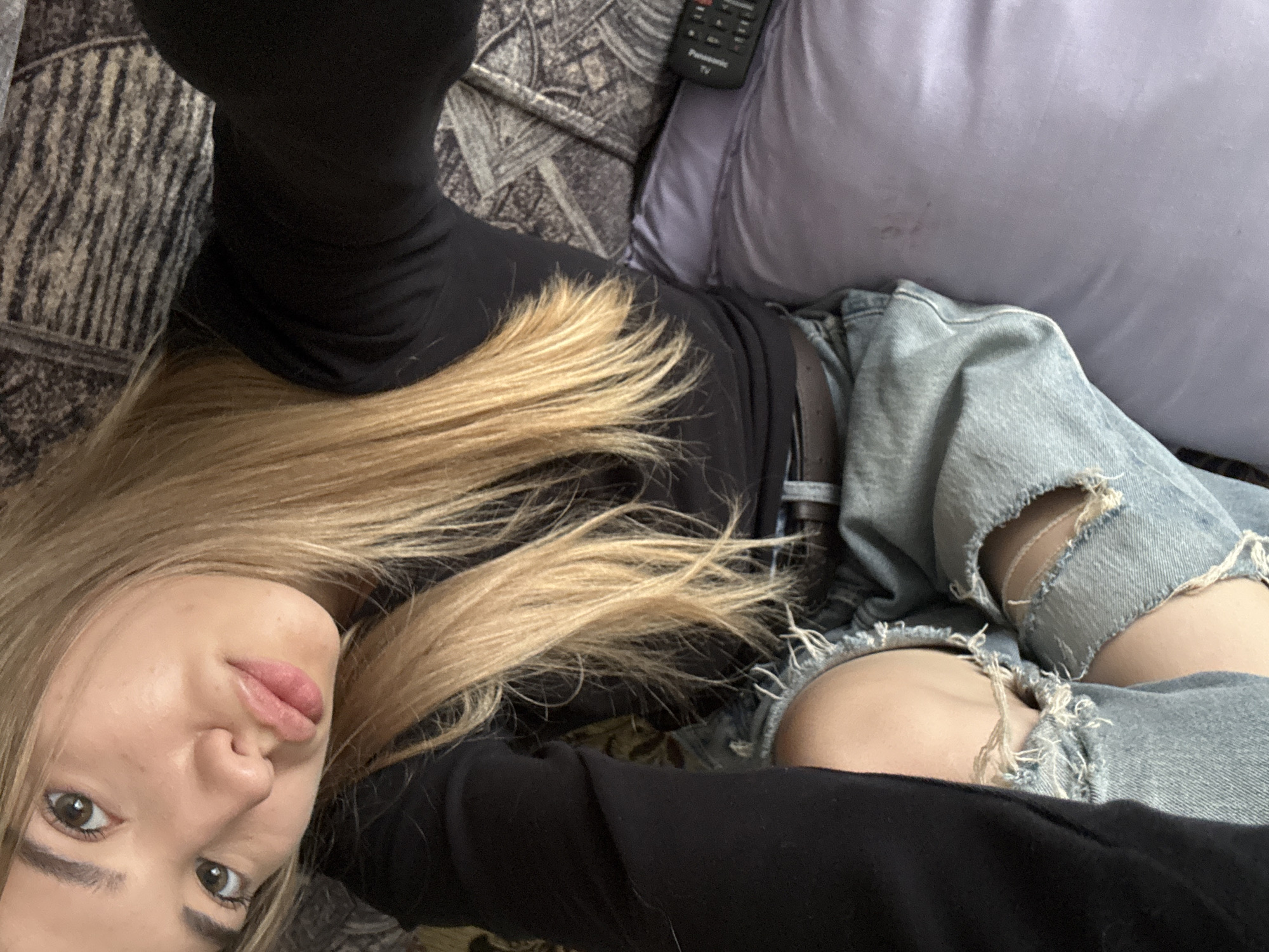 The image shows a young Caucasian female with blonde hair, wearing a black long-sleeved shirt and ripped blue jeans. She is lying down on what appears to be a couch or bed, with a patterned blanket and a purple pillow visible in the background. A remote control is also on the pillow. The woman is looking at the camera with her lips slightly parted. The scene is indoors, and there is no indication of the specific location within Noviy, Russia.