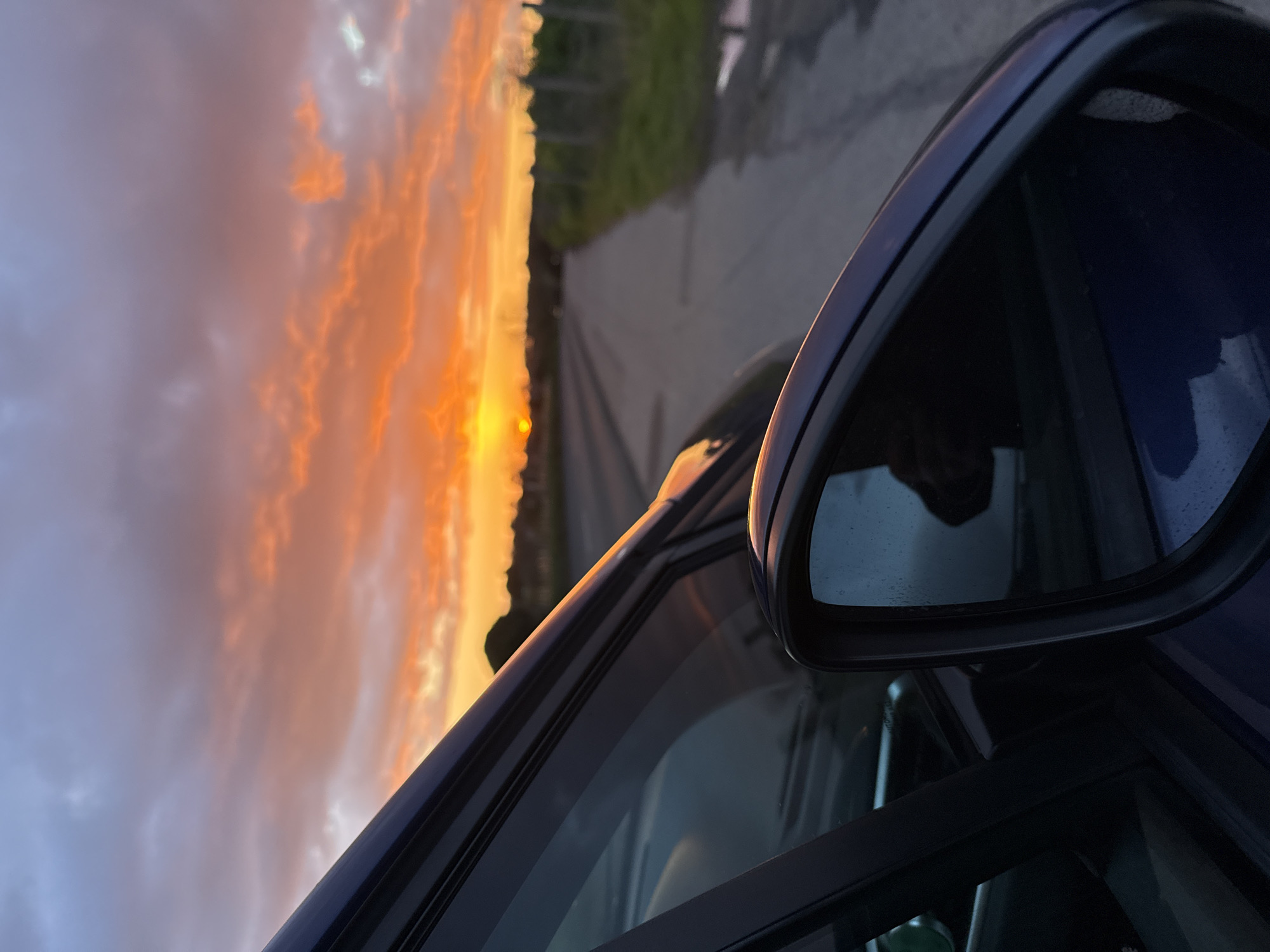 The image, taken from inside a dark blue vehicle, captures a vivid sunset along a road. The sky displays a gradient of colors, ranging from orange and yellow near the horizon to light blue higher up. The sun itself appears as a bright orange orb. A road stretches into the distance, bordered by green foliage. The car's side mirror is prominently positioned in the foreground, partially reflecting the sky. The overall scene appears to be taken in a rural setting. There is no information indicating the location is in Great Yarmouth.