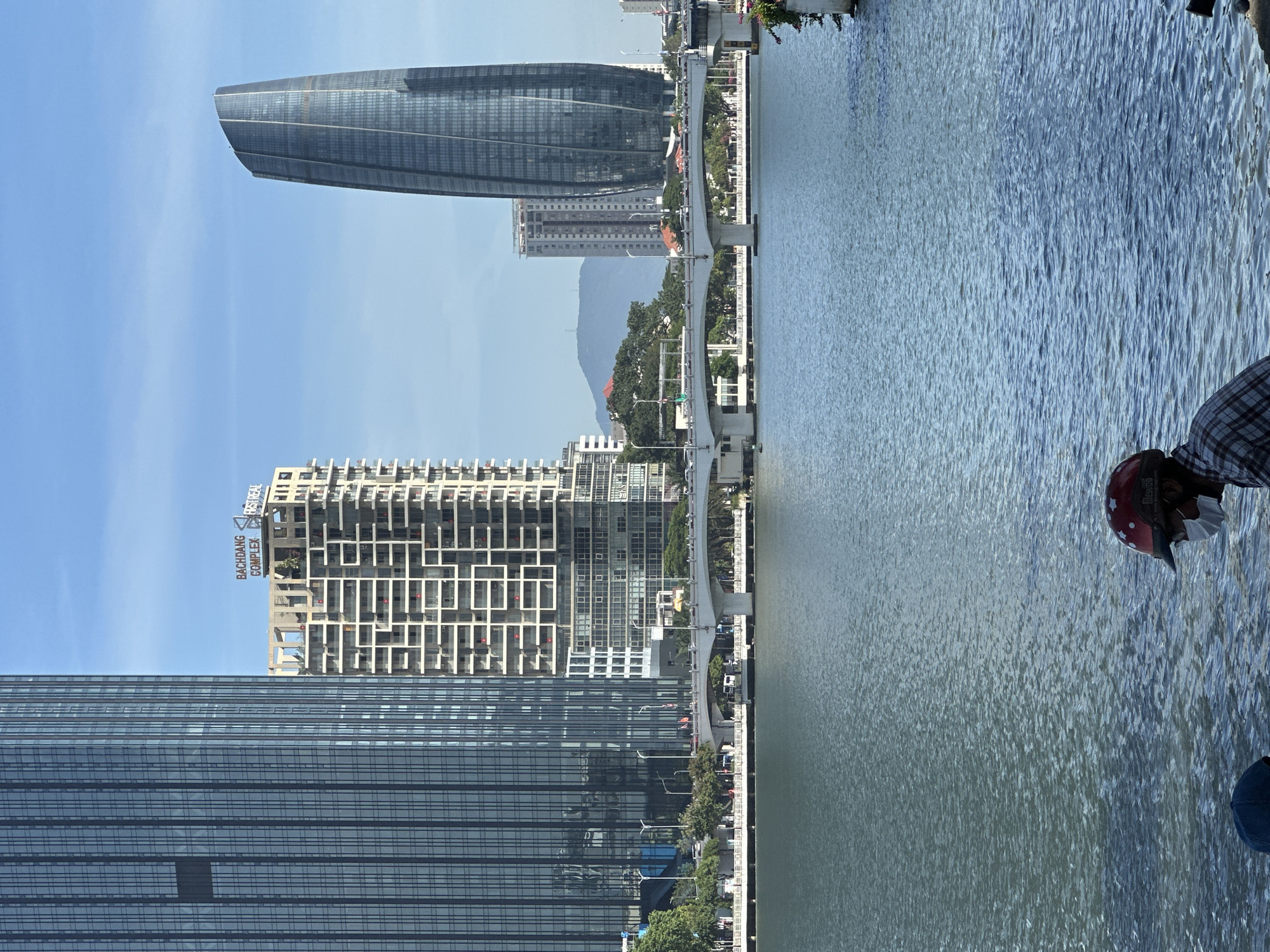The image shows a wide shot of the Han River in Đà Nẵng, Vietnam, with the city skyline in the background. Dominating the left side of the frame is a tall, modern skyscraper with a grid-like facade. To its right, a lighter-colored building with a distinctive patterned exterior is visible, bearing the signage "BAGHDANG COMPLEX FIRST REAL". Further to the right, a curved, modern skyscraper stands prominently against a clear blue sky. A white bridge spans the river, with traffic and trees lining its banks. The foreground is dominated by the shimmering surface of the Han River. In the lower right corner, a person wearing a red helmet with white stars and a plaid shirt is partially visible, facing away from the camera. Another person, wearing a blue cap, is partially visible in the lower left corner. The overall scene depicts an urban waterfront on a bright day.