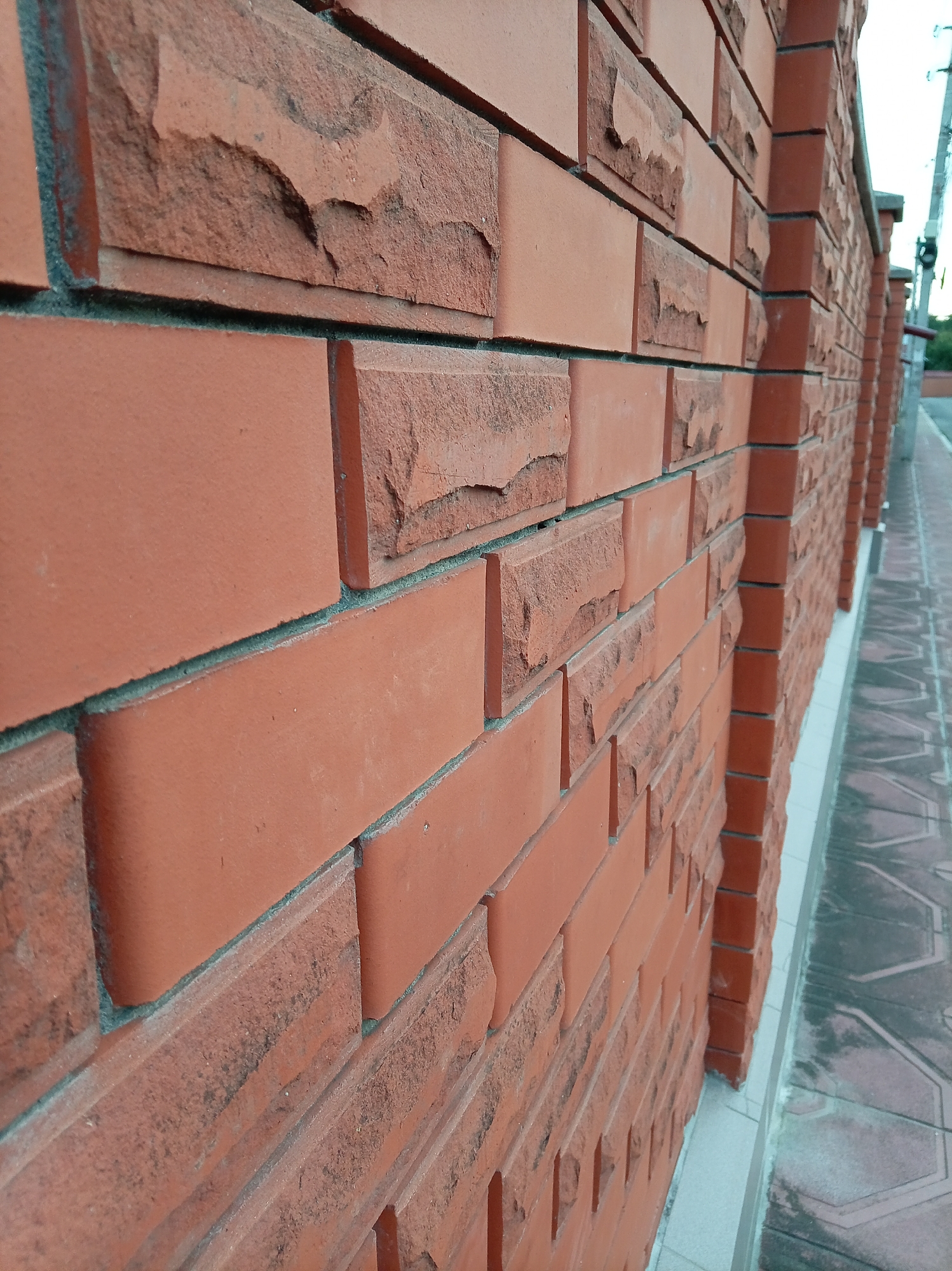 The image shows a close-up of a red brick wall with a sidewalk running alongside it. The wall is constructed with two different types of bricks: some are smooth and uniformly colored, while others have a rough, textured surface with visible imperfections and chipping. The mortar between the bricks appears to be a light gray color. The perspective of the shot emphasizes the texture and depth of the brickwork. In the background, a power line pole and some greenery are visible, suggesting an outdoor urban or suburban setting. The sidewalk is made of patterned paving stones, some of which appear worn or stained. The image is taken from a low angle, looking up at the wall. There are no people or animals present in the image. The lighting suggests it is daytime, possibly overcast. The location is Yessentuki, Russia.