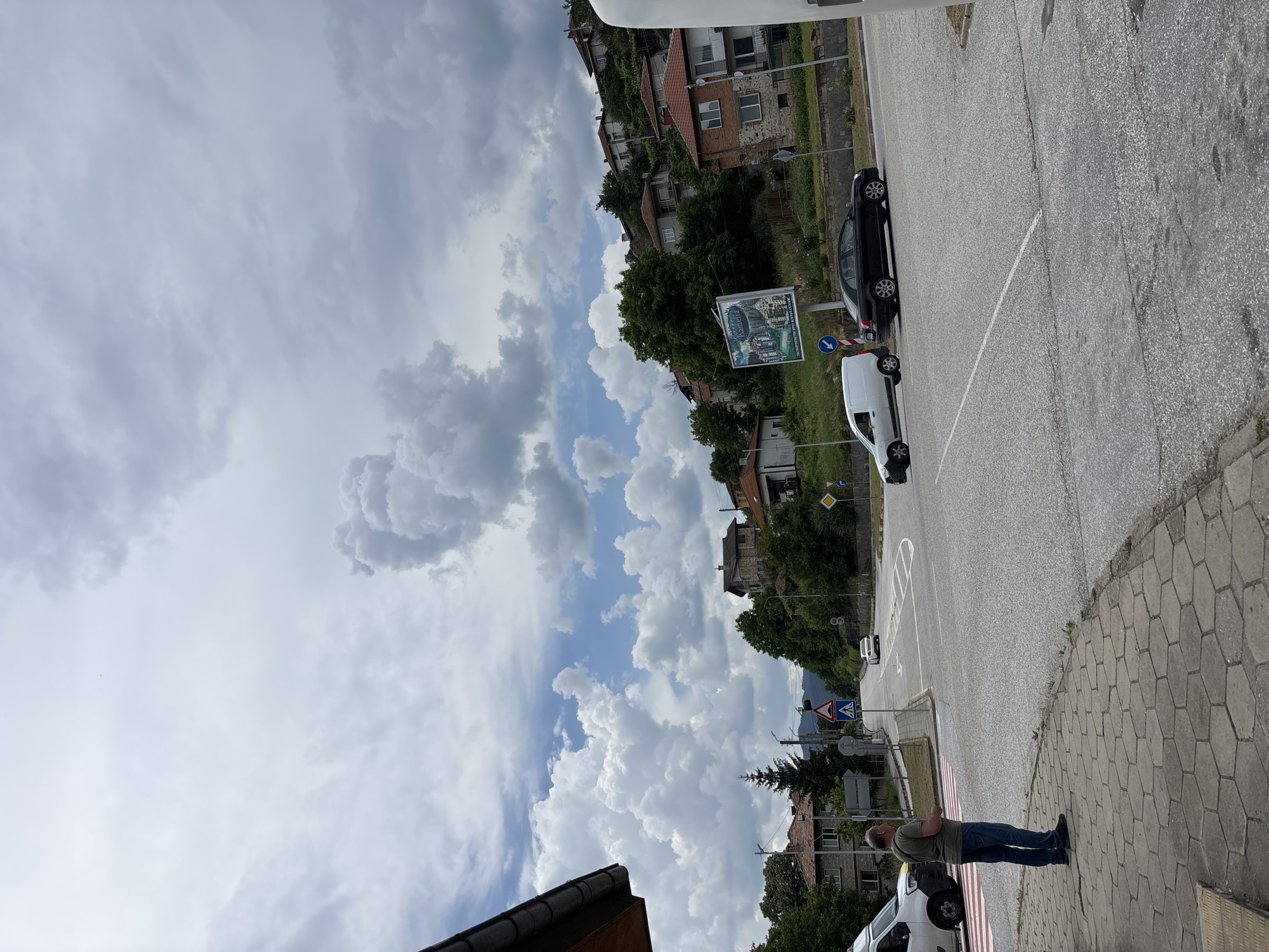 The image depicts a street scene in Hvoina, Bulgaria, under a dramatic, cloudy sky. The perspective is rotated, showing a road running diagonally through the frame, bordered by a sidewalk made of hexagonal pavers. On the sidewalk, a man in a green t-shirt and blue jeans stands with his arms crossed, looking towards the road. A white van is parked on the street next to him.
Further down the road, a black car is parked. Across the street, houses with red tile roofs are nestled into a hillside covered with lush green trees and vegetation. A large billboard with text and an image is visible, advertising a place called "Kamena". Traffic signs, including a blue circular sign with a white arrow and a yellow diamond-shaped warning sign, are present along the roadside.
The weather appears to be overcast, with significant cloud formations filling the sky, suggesting it might be late afternoon or early evening, or that a storm is approaching. The overall atmosphere is quiet and residential, with a hint of a rural or small-town setting given the houses and natural surroundings. The presence of a pedestrian crossing marked by red and white stripes adds to the functionality of the street.