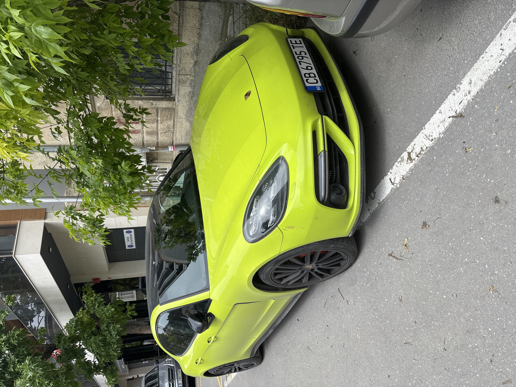 A brightly colored neon yellow (or lime green) Porsche Panamera is parked on an urban street during the daytime. The car is positioned at an angle, with its front facing slightly towards the right and a silver/grey car parked ahead of it. The setting appears to be a city environment, likely in Bulgaria, as indicated by the "BG" on the front license plate.
The Porsche features black multi-spoke wheels with red brake calipers visible behind them. Its front bumper shows some minor scuff marks near the right headlight. The license plate clearly reads "CB 6795 TE".
To the left of the car, green foliage from trees partially obscures a light-colored building with a window protected by an ornate black metal grille. A downspout is also visible on this building. To the right of the car is an asphalt road surface with a clear white painted line, suggesting a parking space or lane marker. Small pieces of natural debris, like leaves and twigs, are scattered on the asphalt.
In the background, behind the Porsche, more buildings are visible. One prominent building has a modern design with large glass panels. On its entrance, a sign with Cyrillic text is discernable, reading "ОДИТ И КОНСУЛТАНТСКА" (meaning "Audit and Consulting" in Bulgarian), accompanied by two circular logos, possibly national or organizational emblems. The overall lighting suggests it's not direct harsh sunlight, perhaps an overcast day or early morning/late afternoon. No people are visible in the image.