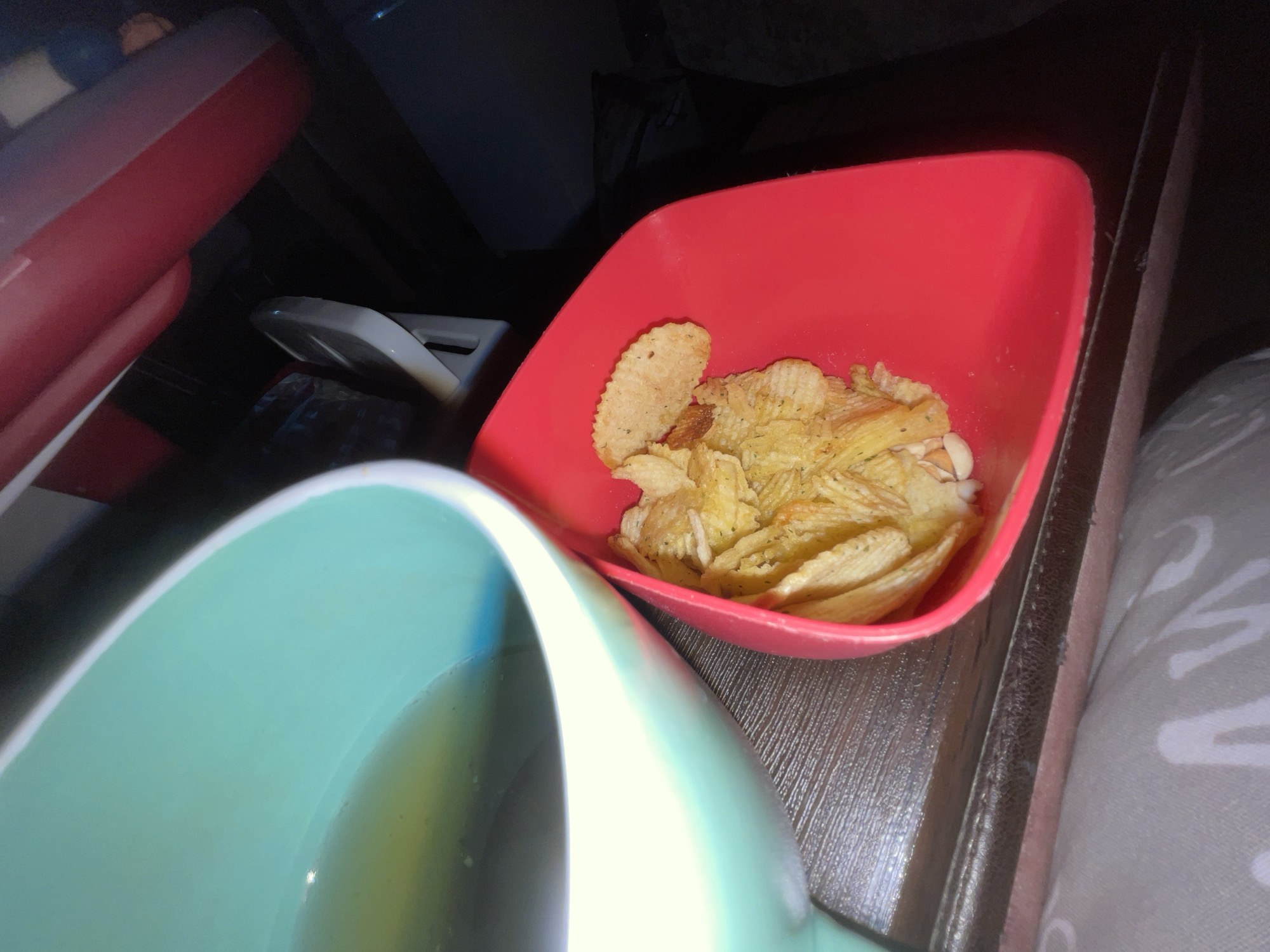 A close-up shot depicts a partially eaten bowl of ruffled potato chips in a red, square plastic bowl, accompanied by a blue mug filled with liquid. The scene is set on a dark, possibly wooden, surface, with the edge of a patterned cushion or blanket visible on the right. A plastic container is visible in the top left and a black object in the top center. The location cannot be determined with the available information.
