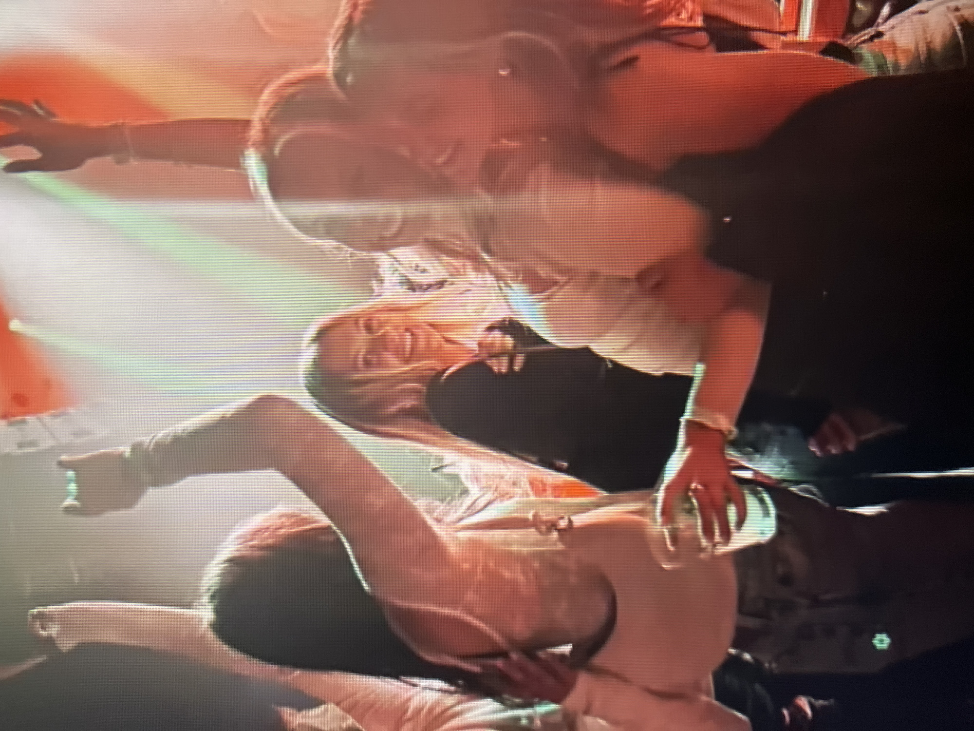 In a dimly lit, smoky environment with vibrant red and green lights illuminating the scene, several young women are seen dancing and enjoying themselves. The image appears to be taken at a concert or club. One woman in the foreground, wearing a white top and jeans, is holding a drink and smiling. She is surrounded by friends, with one woman embracing her from behind. Other women with raised hands suggest an energetic atmosphere, possibly during a performance. The overall impression is of a lively social gathering.